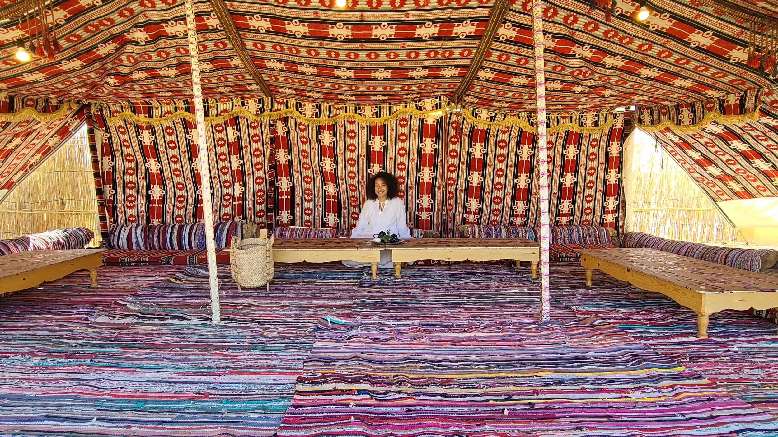 A woman sitting in a traditional colorful tent with patterned fabric walls and ceiling, wooden furniture, and woven rugs on the floor.