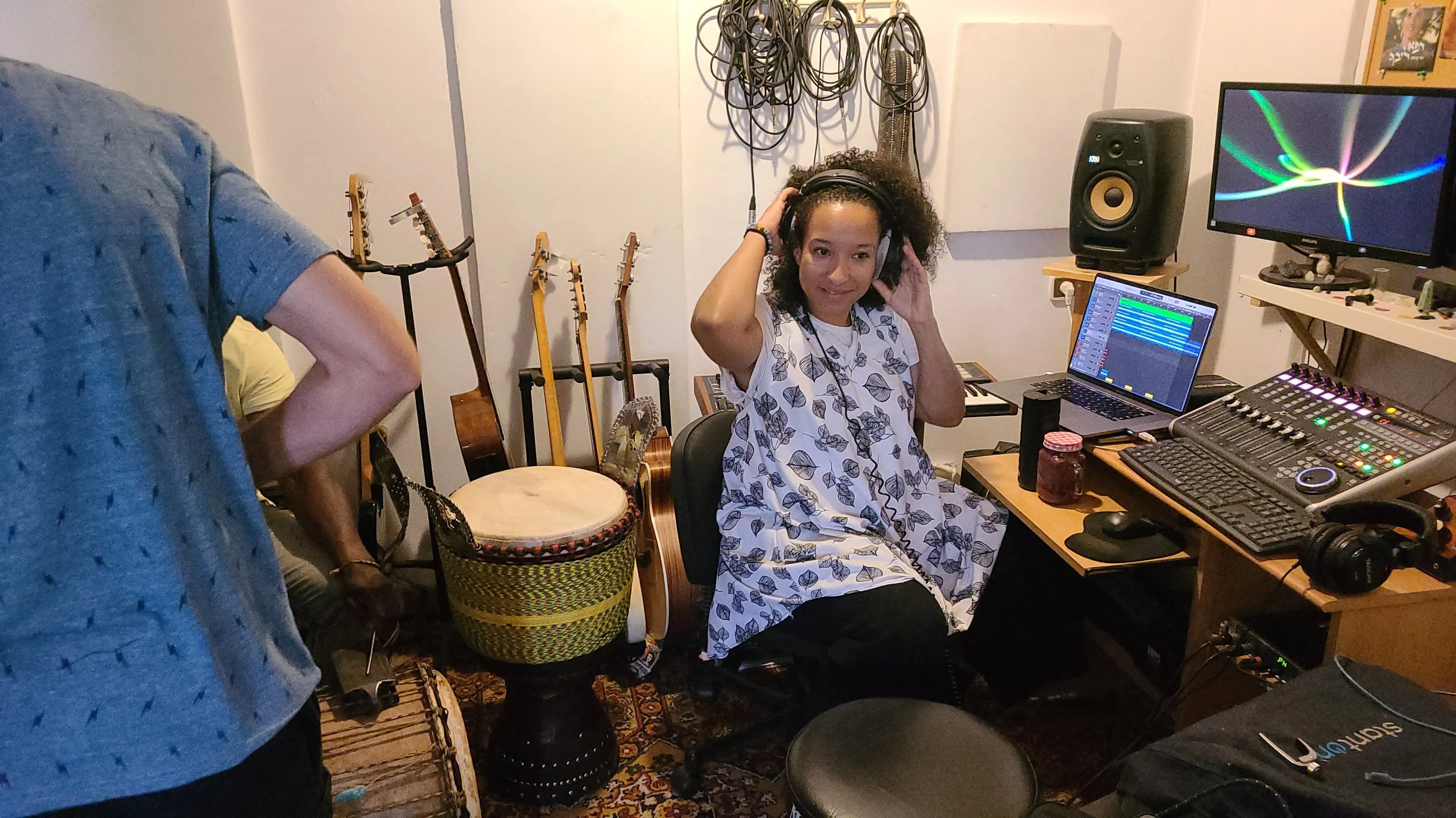 A woman with curly hair wearing headphones, sitting in a music studio surrounded by musical instruments, audio equipment, and computer monitors.