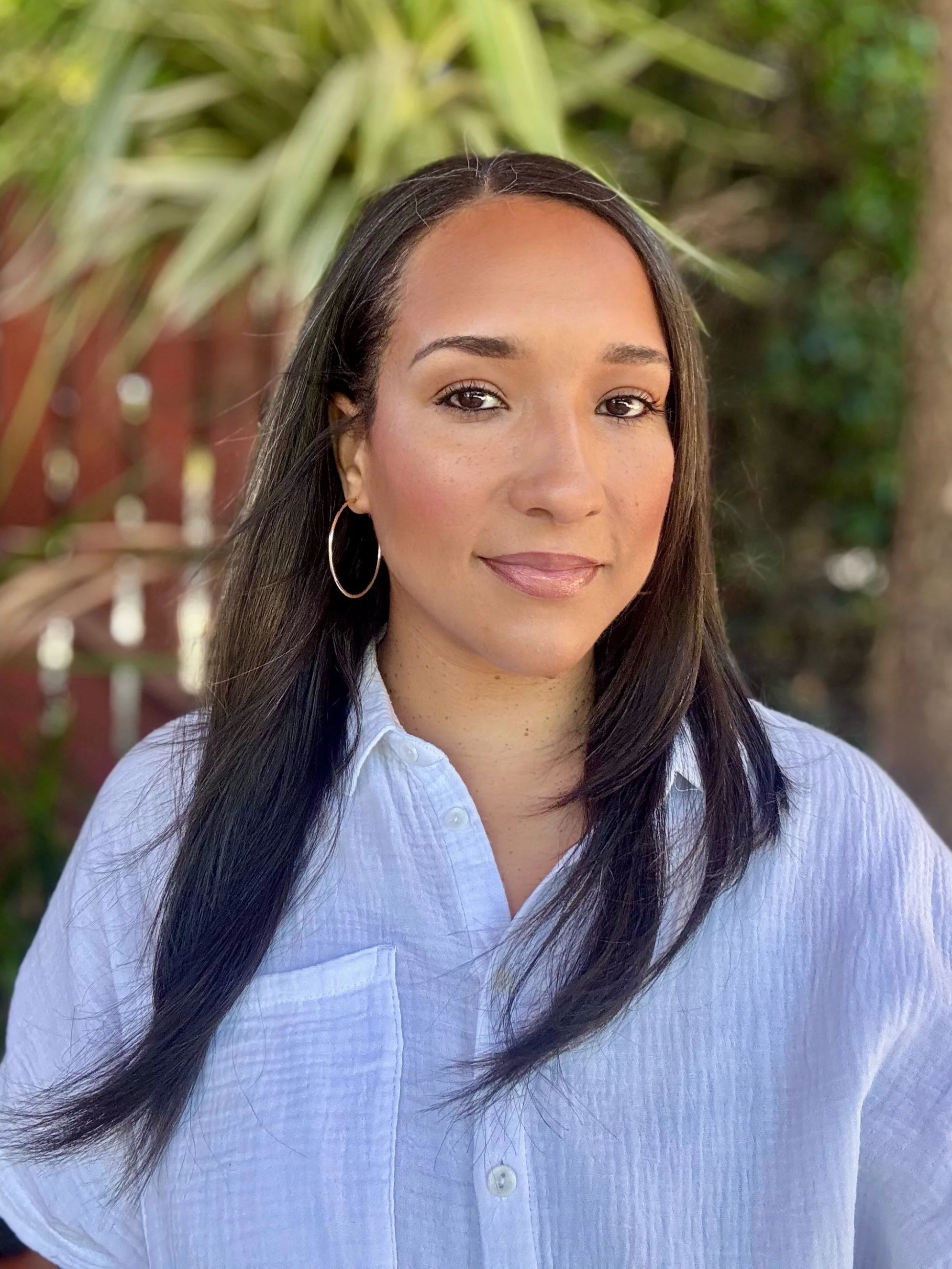 Close-up of a woman with long dark hair, hoop earrings, and light makeup, wearing a white button-up shirt, standing outdoors with green foliage in the background.