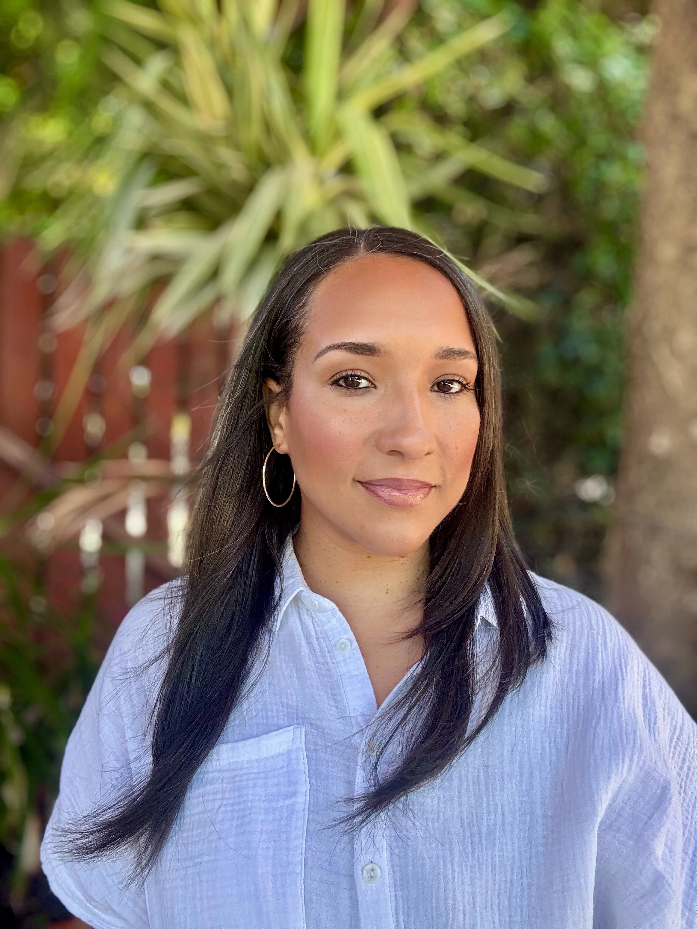 Close-up of a woman with dark hair wearing large hoop earrings and a white button-up shirt, standing outdoors with green foliage and a tree in the background.