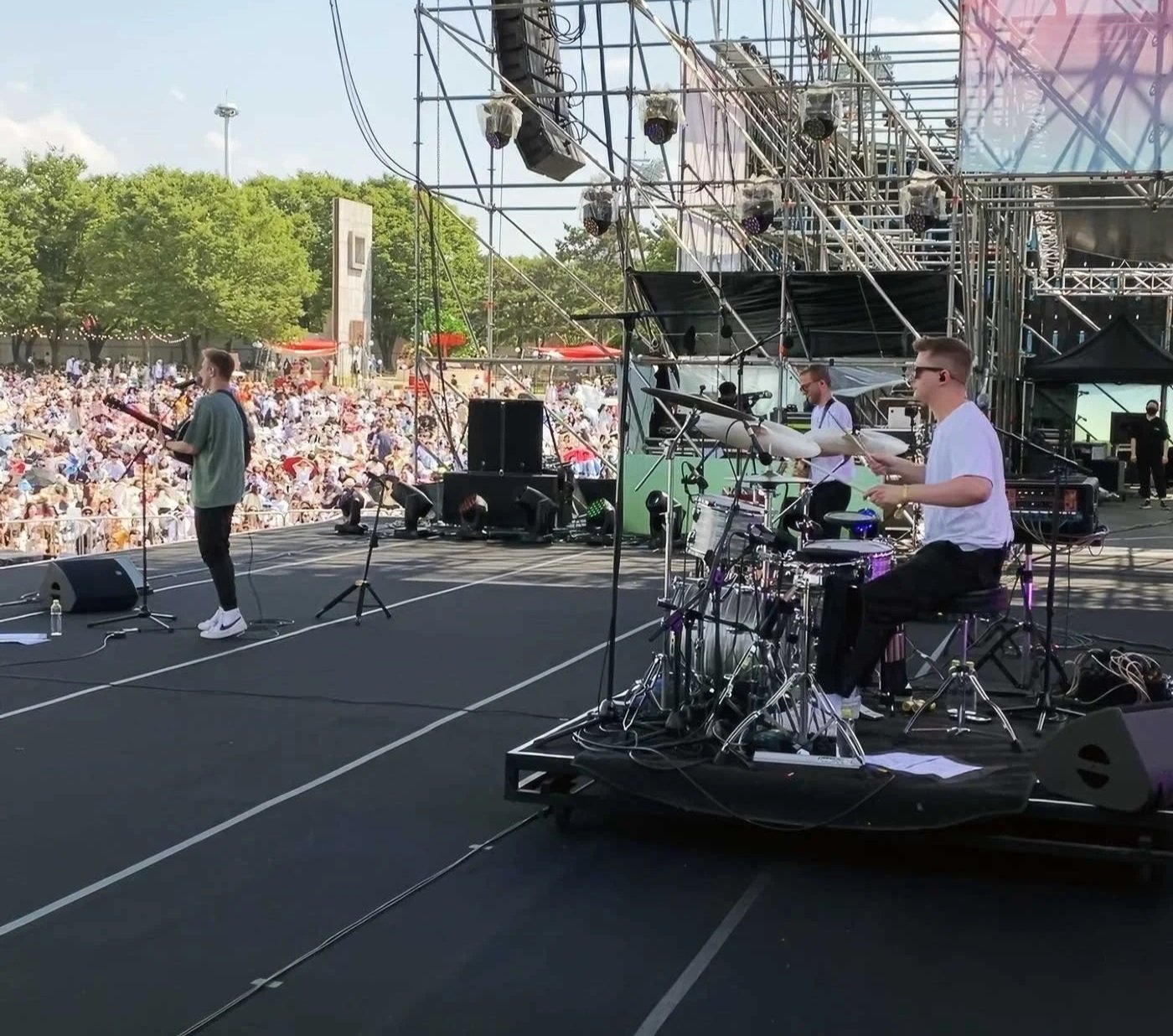 A band performs on an outdoor stage during a concert, with a large crowd in the background. The stage has musical instruments and scaffolding, and the sky is clear with some trees visible.