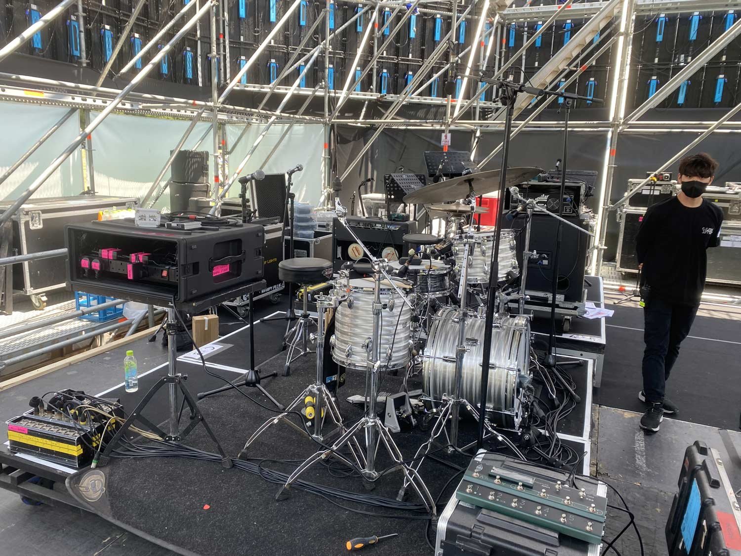 Music stage setup with drums, sound equipment, and microphone stands at an outdoor venue, with a person wearing a black mask and black clothing standing nearby.