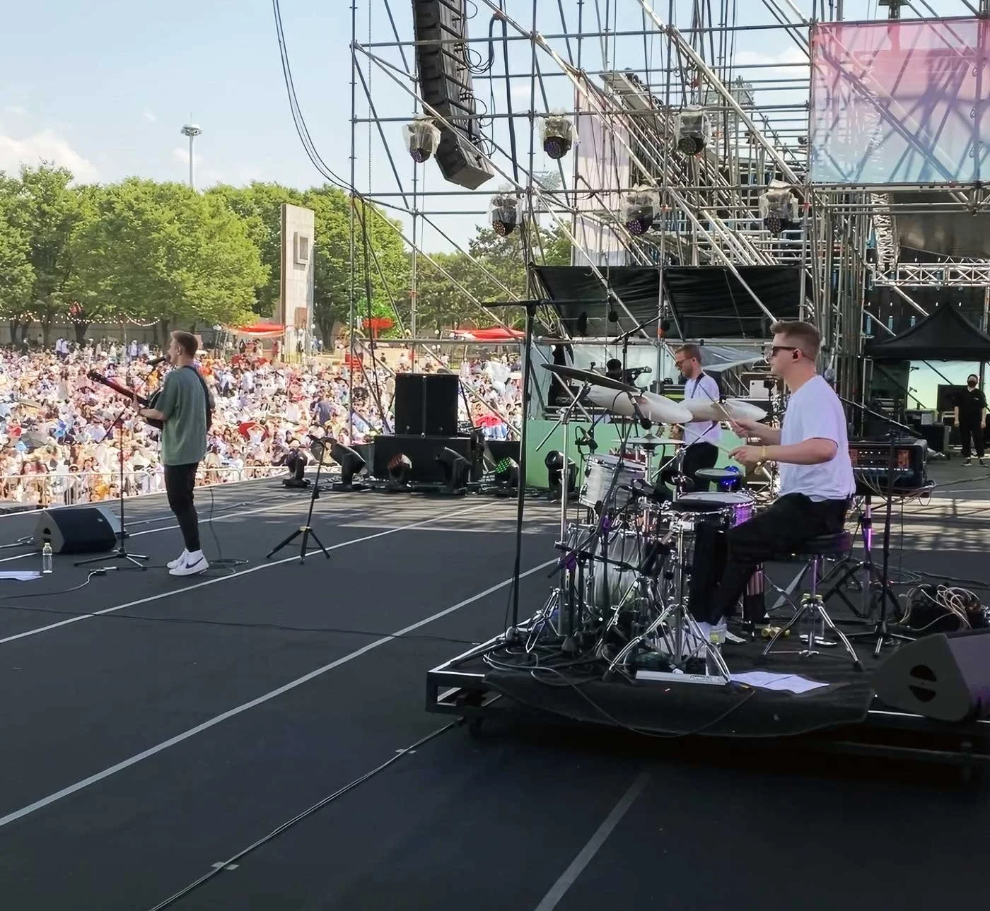 A band performing live on an outdoor stage in front of a large audience during daytime. The stage has musical instruments and sound equipment with trees and a crowd of spectators in the background.