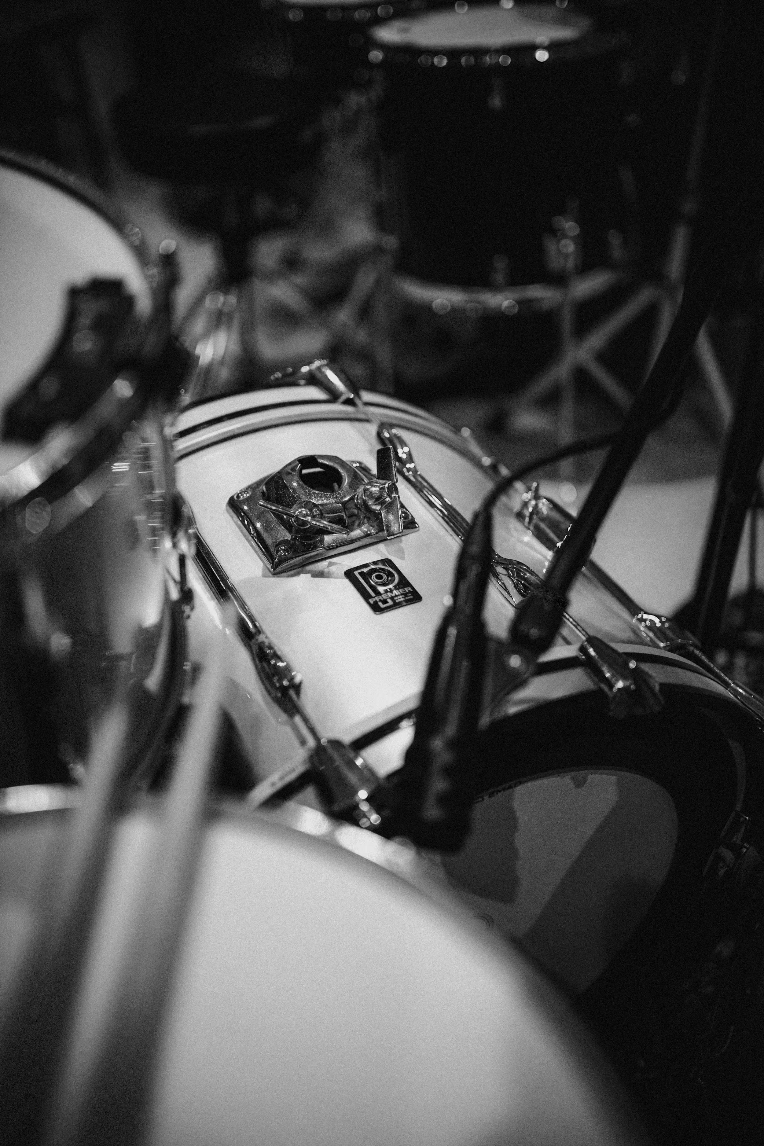Close-up black and white photo of a vintage drum set, showing part of a bass drum with a club logo and a hardware mount, with some drumsticks visible and a blurred background.