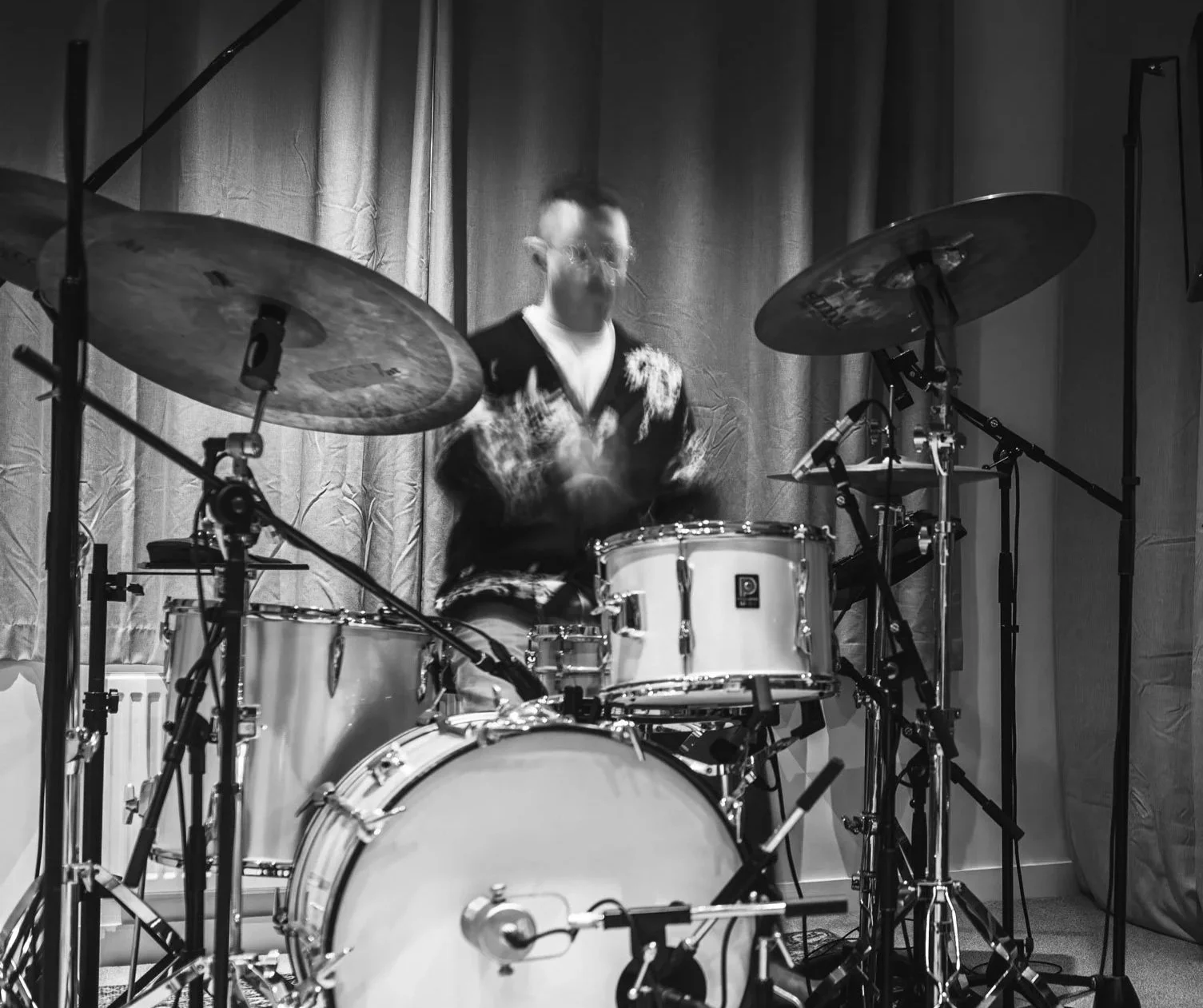Black and white photo of a man playing drums on stage with curtains in the background.