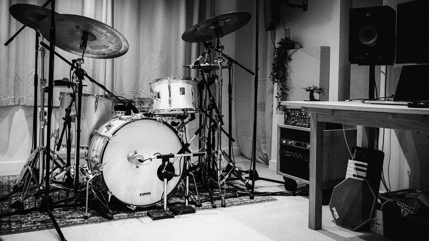 A drum set with cymbals and a bass drum on a rug in a recording studio or rehearsal space, with music equipment and a laptop on a wooden desk nearby.
