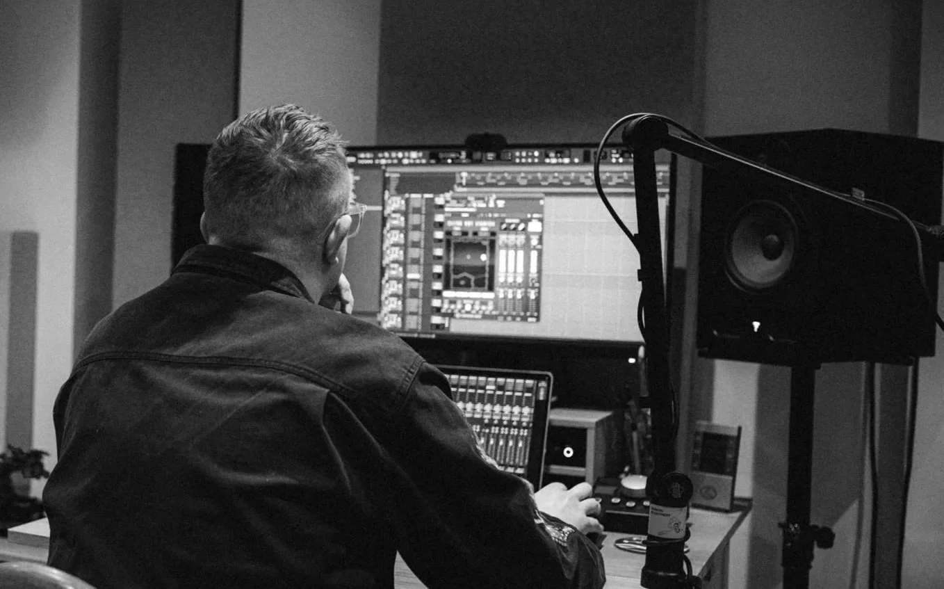 A person wearing glasses and a black jacket working in a music studio, looking at audio editing software on a large computer screen with studio speakers nearby.