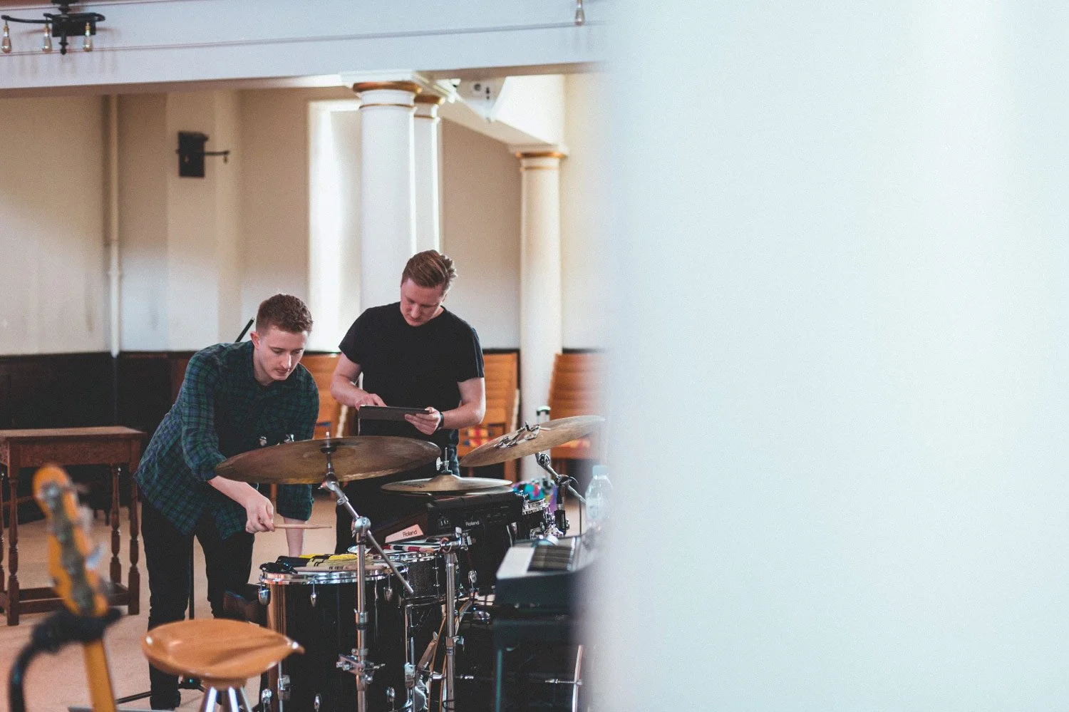 Two young men tuning a drum set in a music rehearsal space.
