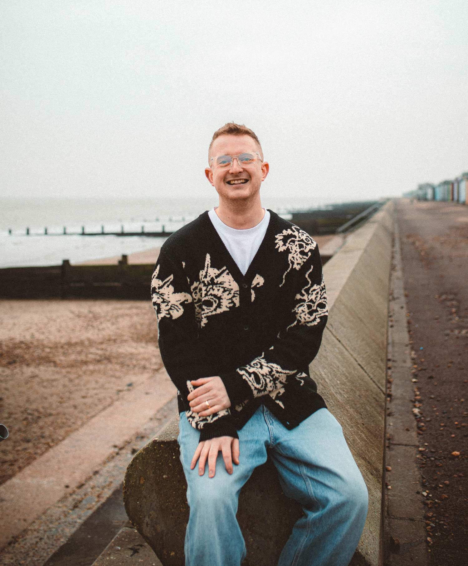 A smiling man with glasses and short hair, wearing a black and white patterned sweater and light blue jeans, sitting on a rock beside a concrete ledge at the beach during daytime with a cloudy sky.