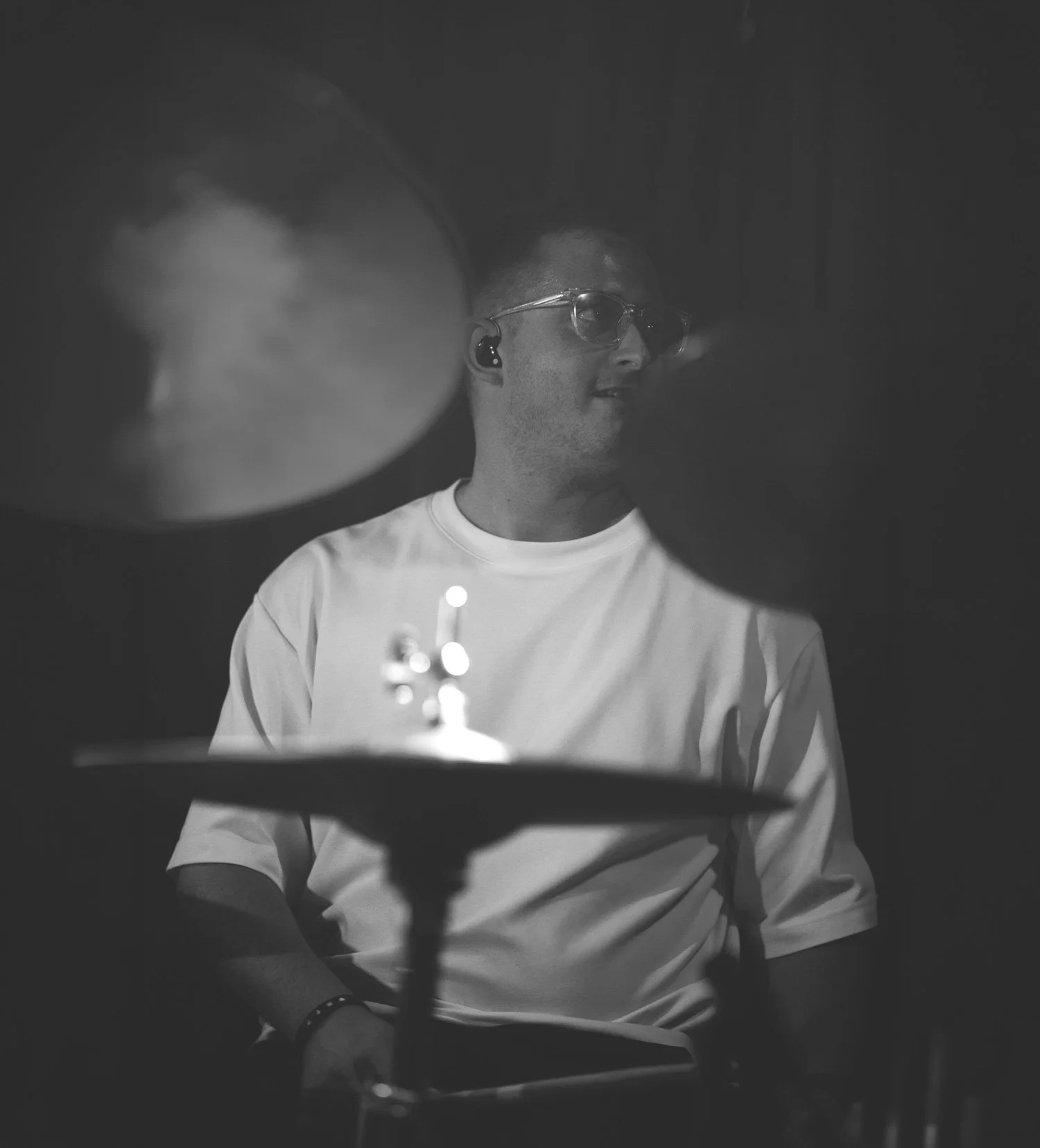Black and white photo of a man wearing glasses and white t-shirt, viewed through a drum cymbal.