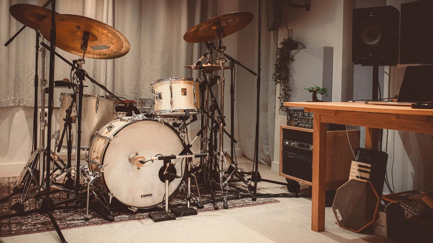 A drum set on a rug in a music studio with audio equipment and a wooden desk with a laptop.