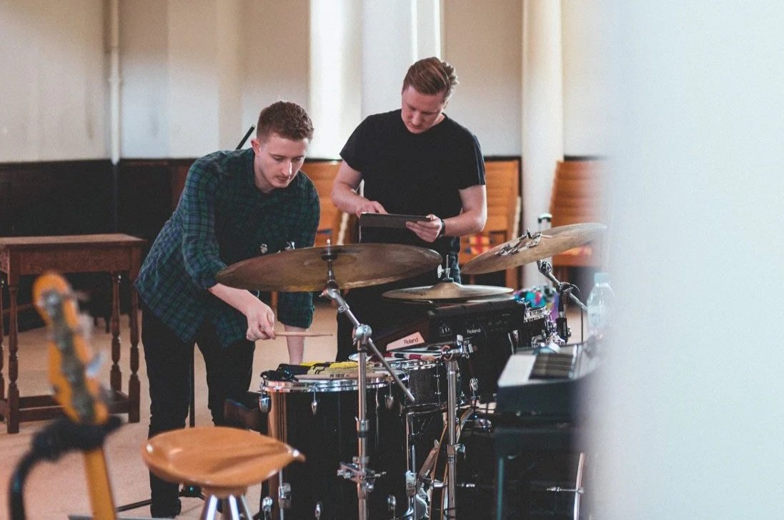 Two young men tuning a digital drum set in a music rehearsal space.