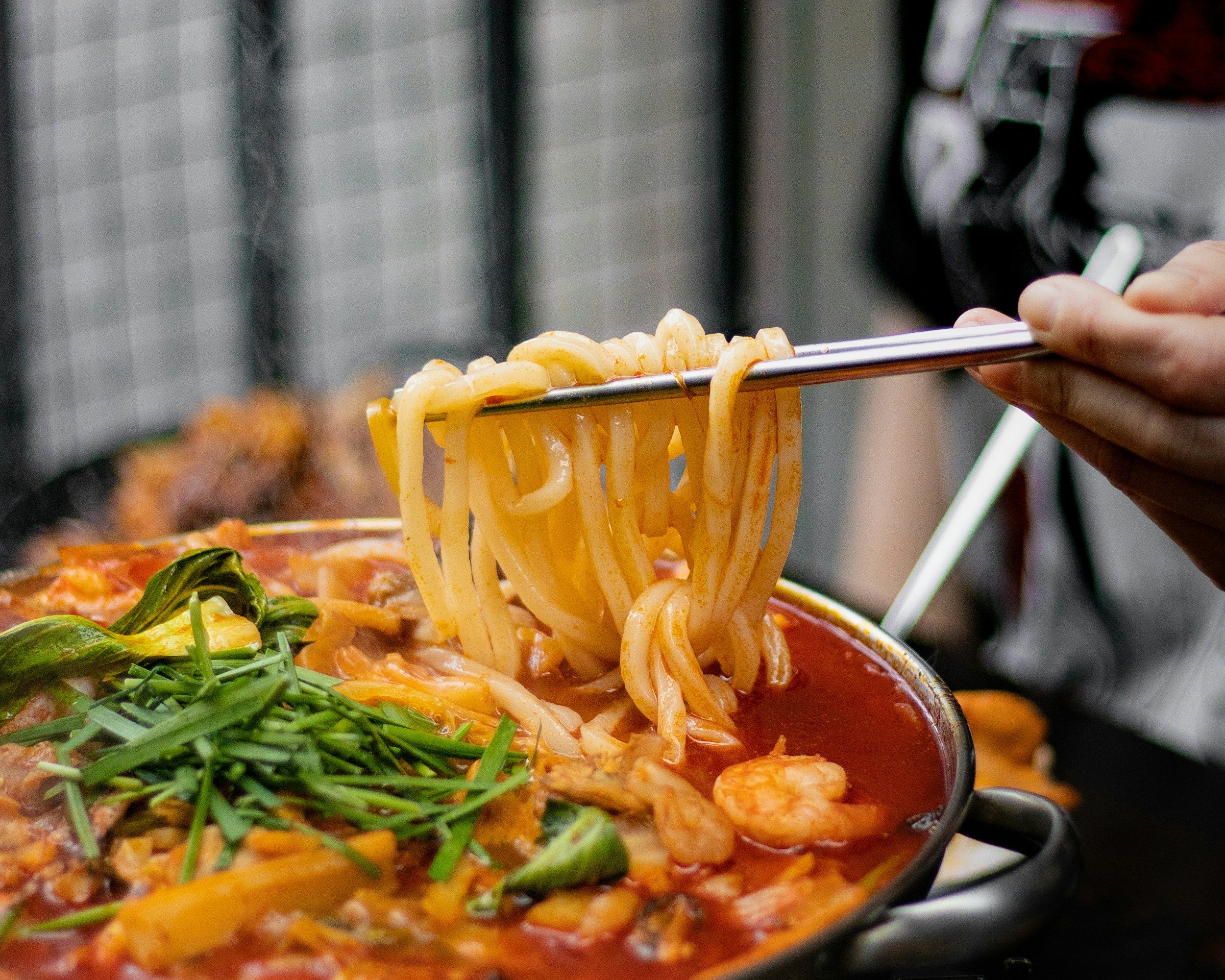 Close-up of a person using tongs to serve spaghetti pasta with sauce into a bowl, with fresh green vegetables visible in the foreground, in a restaurant or kitchen setting.