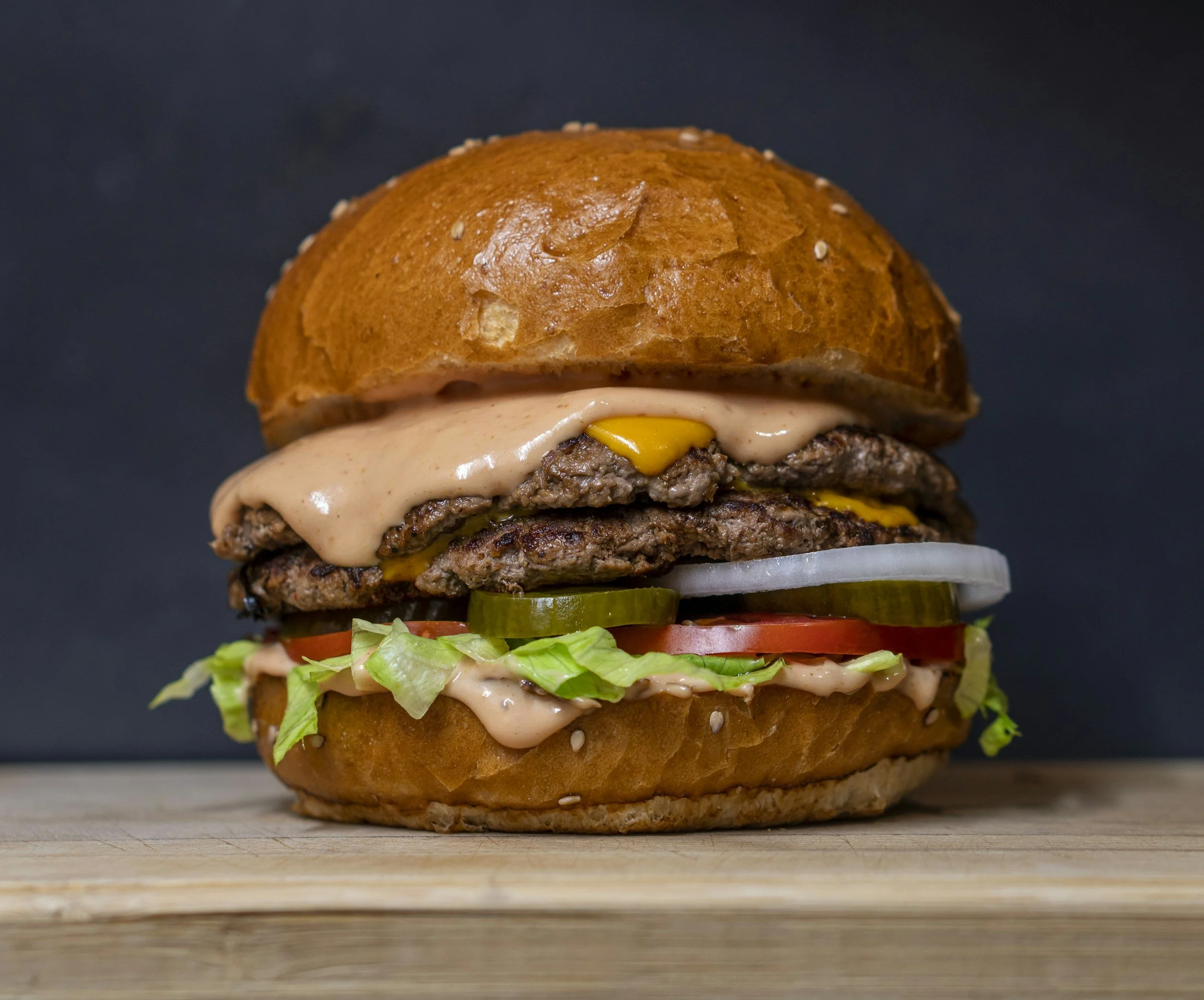 Close-up of a double cheeseburger with lettuce, tomato, pickles, onion, and melted cheese, in a sesame seed bun, on a wooden surface against a black background.