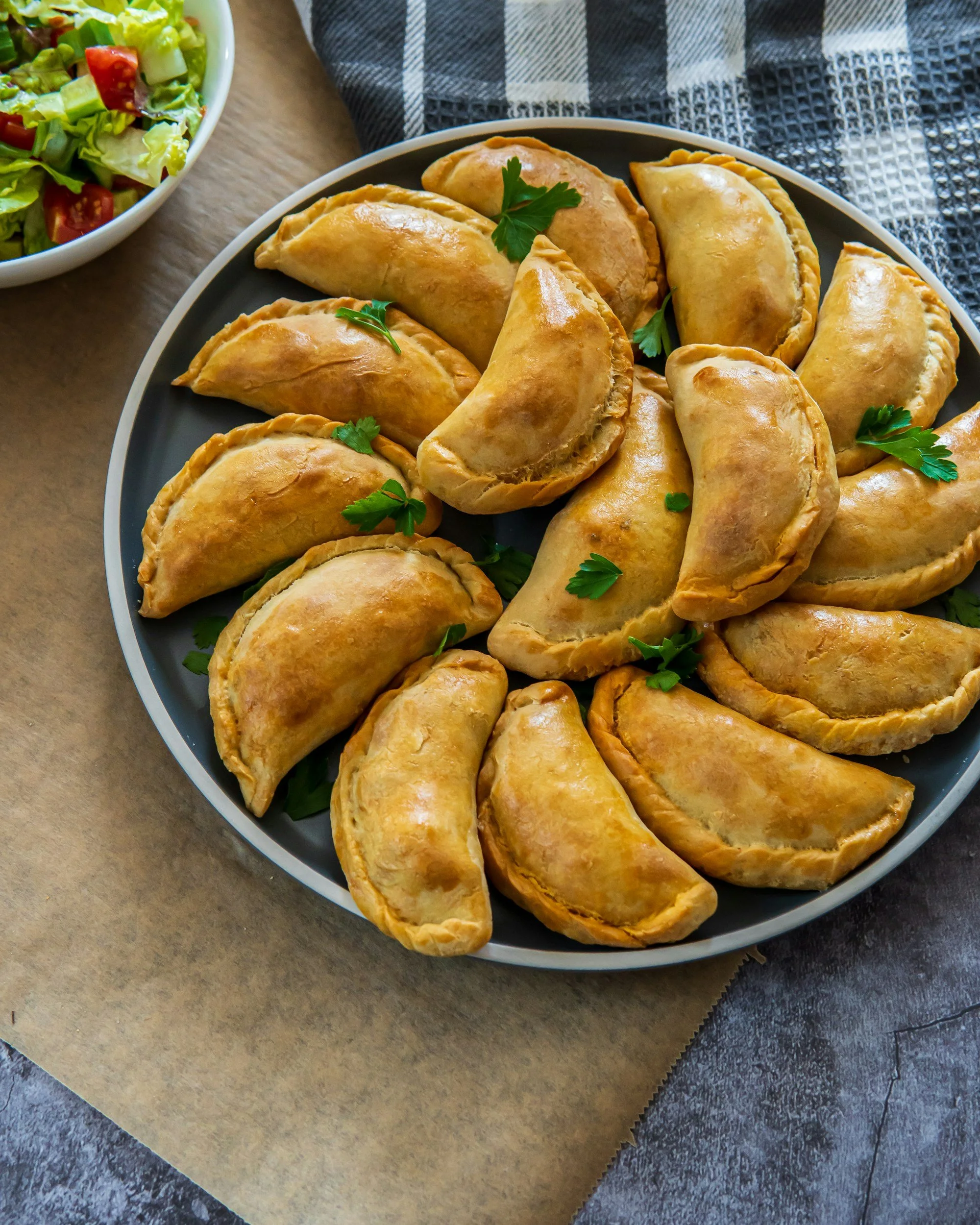 Plate of cooked samosas garnished with fresh parsley, alongside a side salad with lettuce and cherry tomatoes, on a dark surface with black and white checkered cloth.