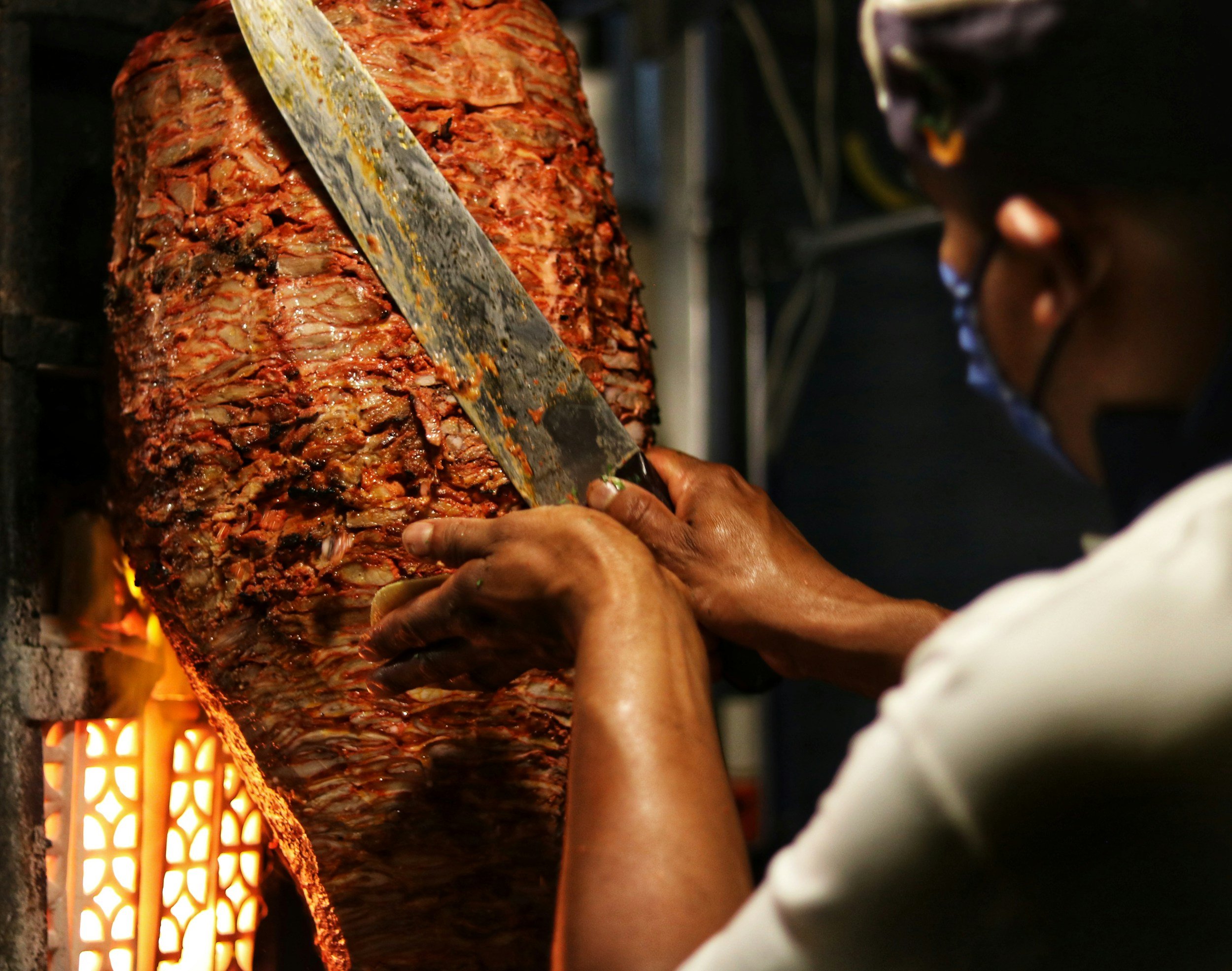 A person is slicing a large, cooked spit of meat, likely shawarma or gyro, with a large knife. The meat is roasting on a vertical rotisserie over an open flame.