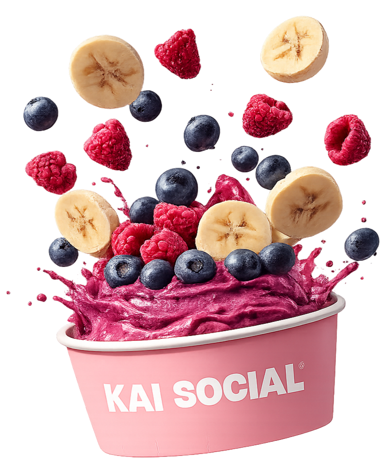A pink bowl labeled "KAI SOCIAL" with purple and pink smoothie topped with sliced bananas, blueberries, raspberries, and some berries are falling into the bowl.