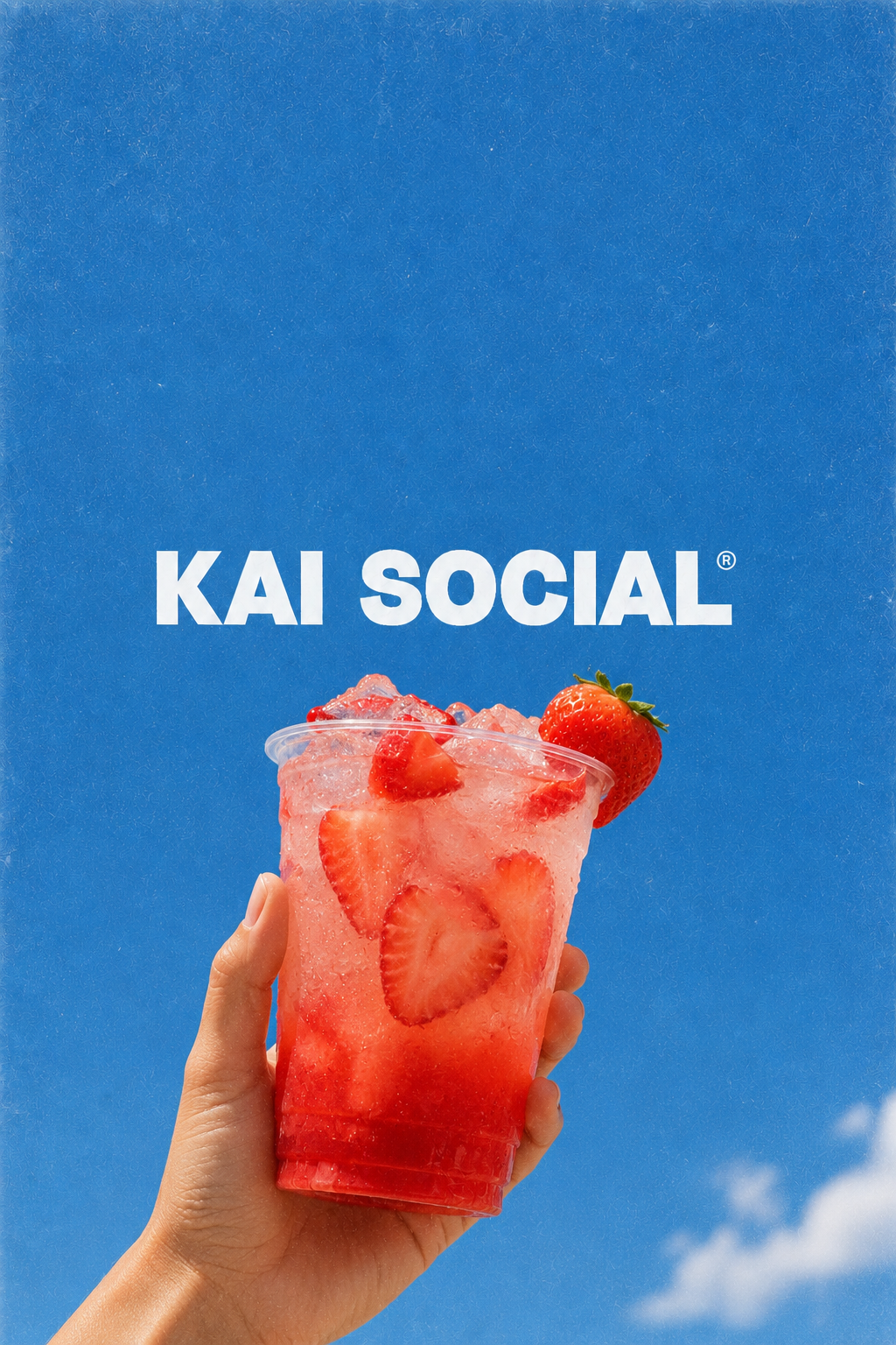 A hand holding a clear plastic cup filled with a pink strawberry drink and ice cubes, with a large strawberry on the rim against a blue background with the text 'KAI SOCIAL'.