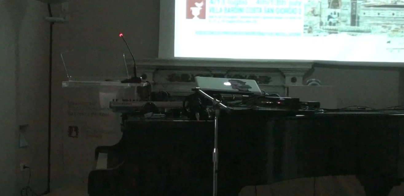 A music setup with a black grand piano, a microphone on a stand, a laptop, and a small transparent box with a microphone and a warning sign, set in a dimly lit room with a projector displaying a presentation or slide show on the wall.