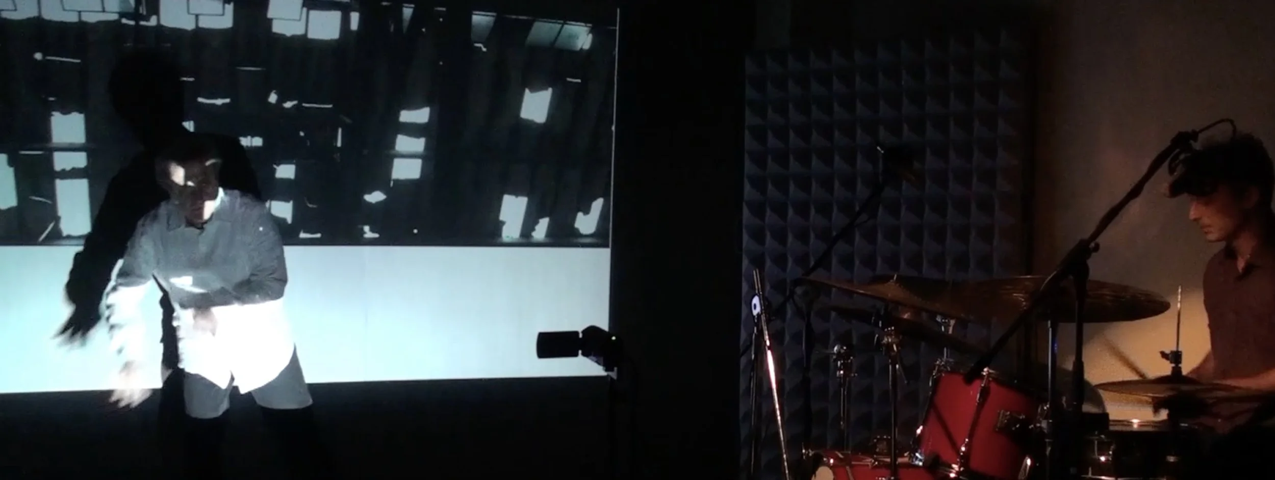 Two musicians performing in a recording studio. One is dancing or moving energetically in front of a large screen, while the other is playing drums wearing headphones.