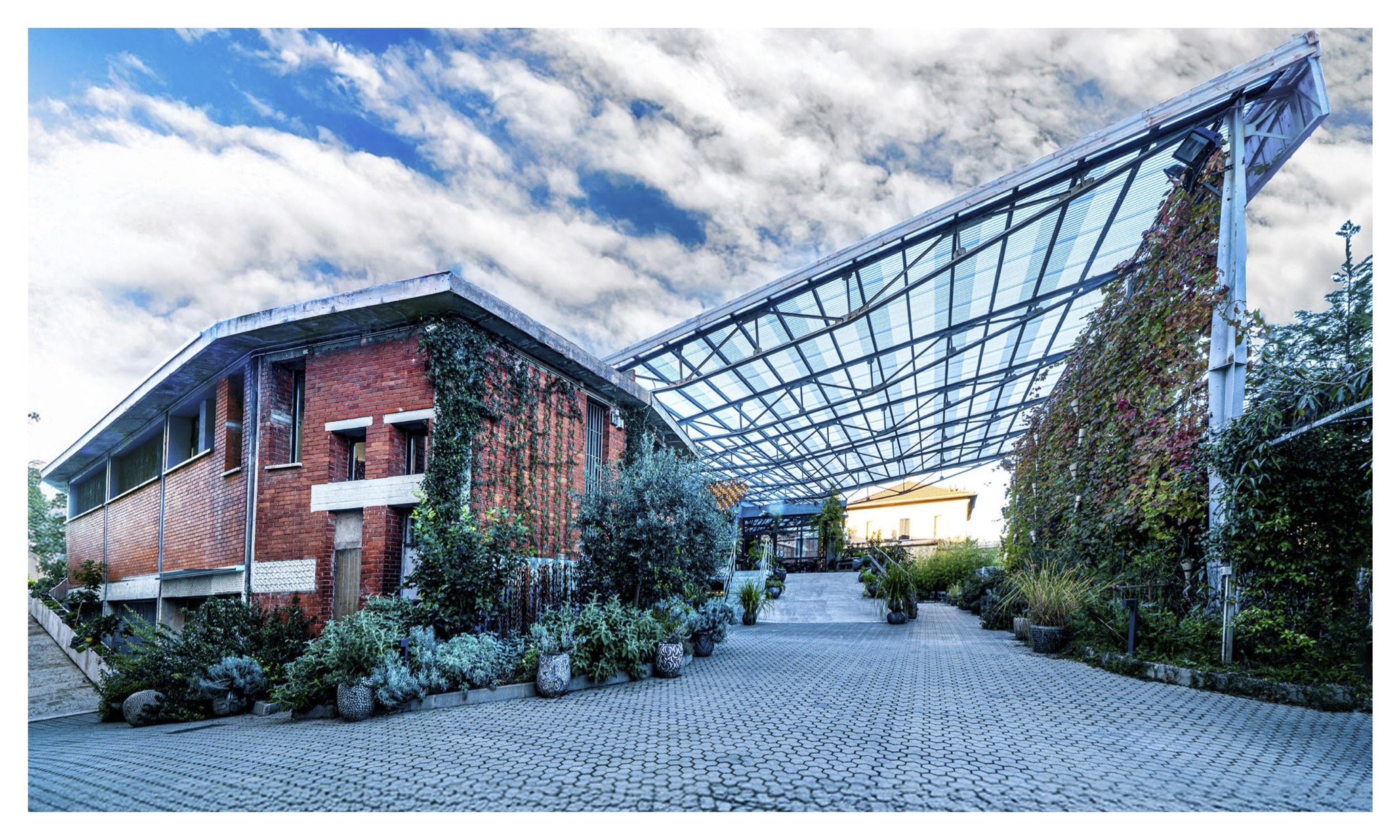 A brick building fondazione suzzani in milan with a curved roof and greenery climbing up its side, next to a glass and metal structure covered with vines, under a cloudy sky.
