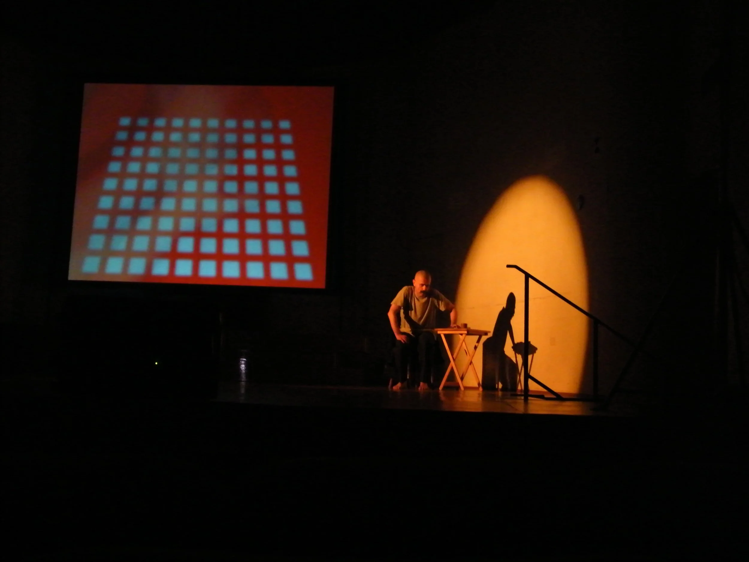 A dark stage with a man seated at a table, a large red screen with a grid pattern projected, and a large illuminated object resembling an egg or bullet in the background.
