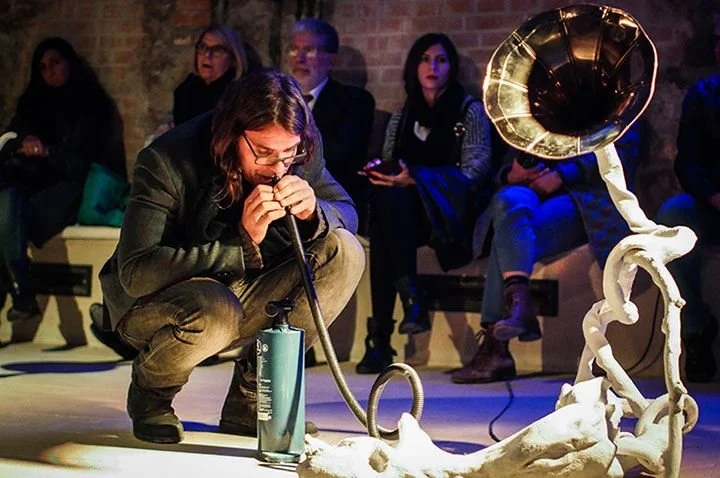 simone pappalardo performing with a musical instrument resembling a gramophone, in front of an audience in a dimly lit venue with brick walls.
