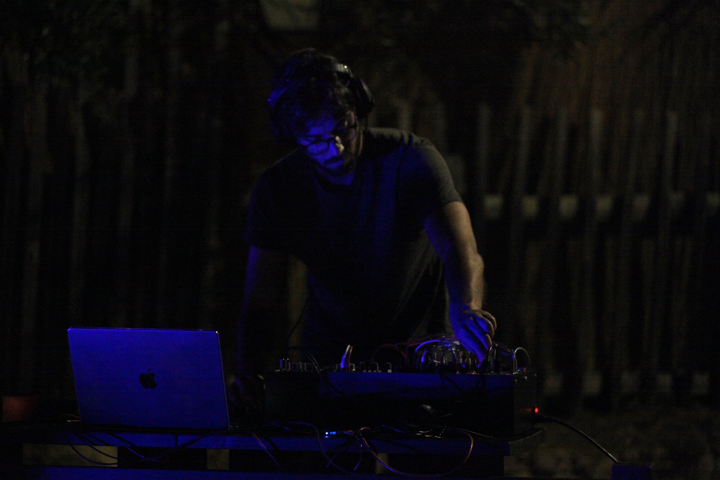 A person with curly hair and glasses wearing a dark shirt, operating DJ equipment with a DJ turntable and a laptop in a dimly lit environment.