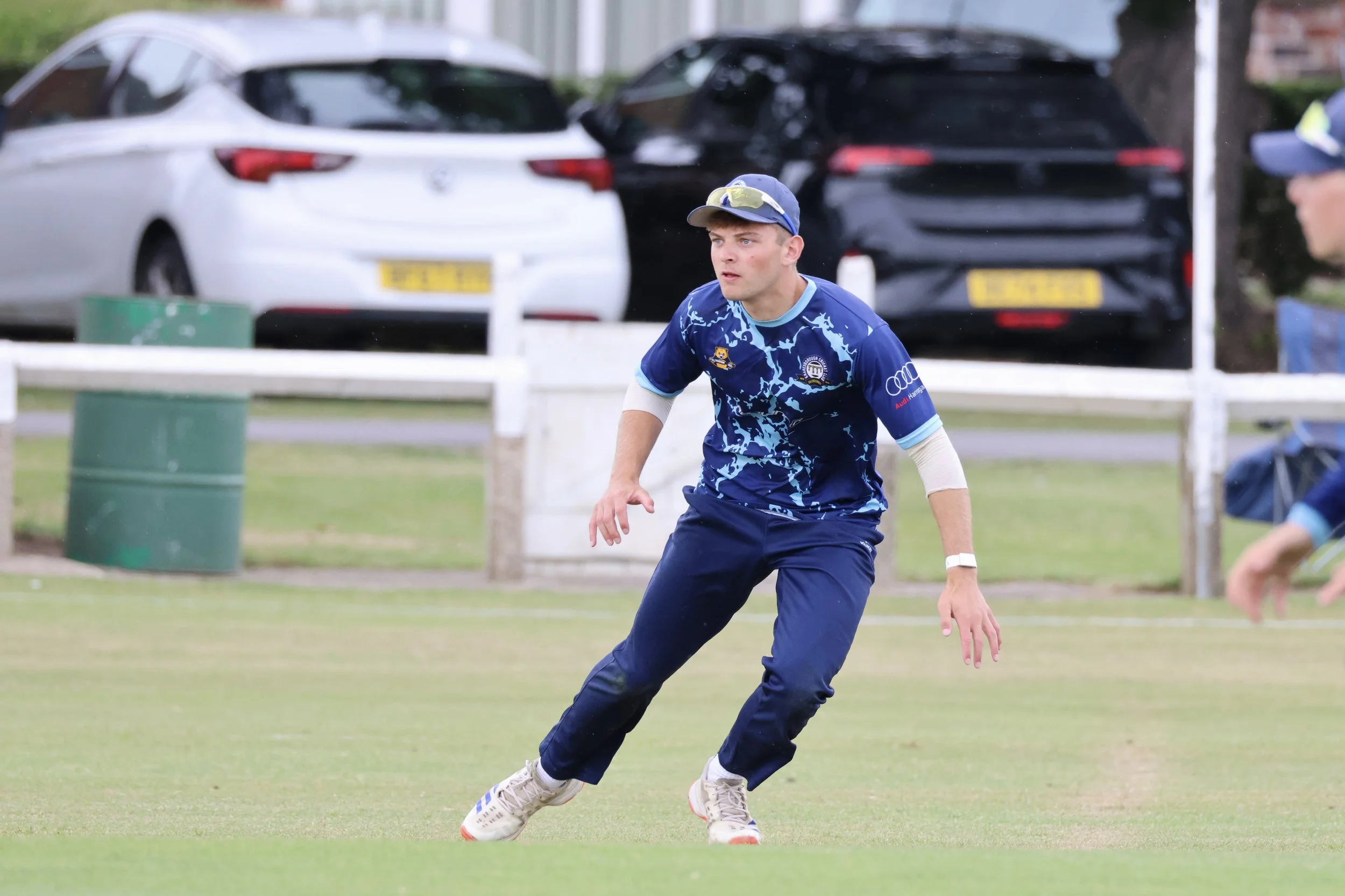 A male athlete wearing a blue sports jersey, white long sleeves, and a baseball cap, playing cricket outdoors on a grassy field with parked cars and a person in the background.
