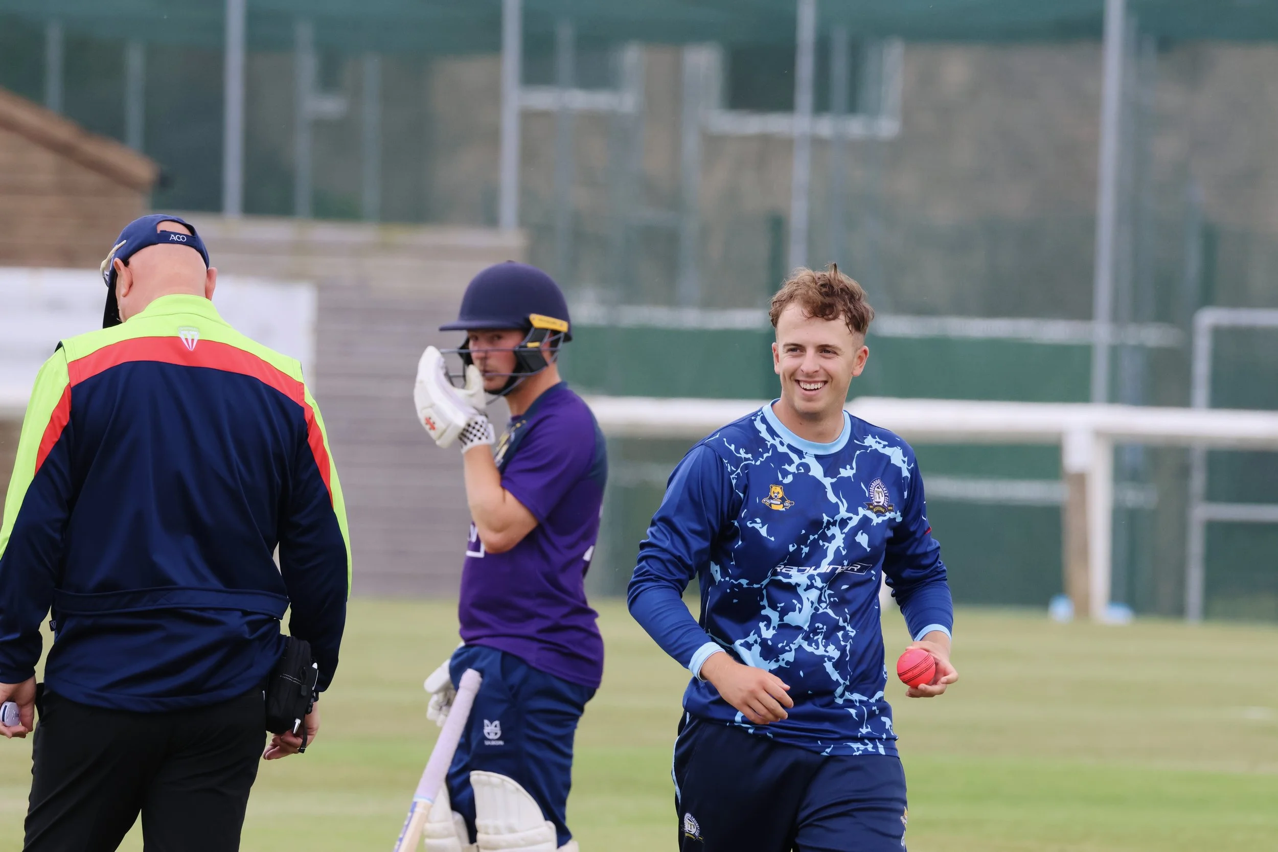 Two men in cricket uniforms smiling and holding a cricket ball while talking to an umpire on a cricket field.