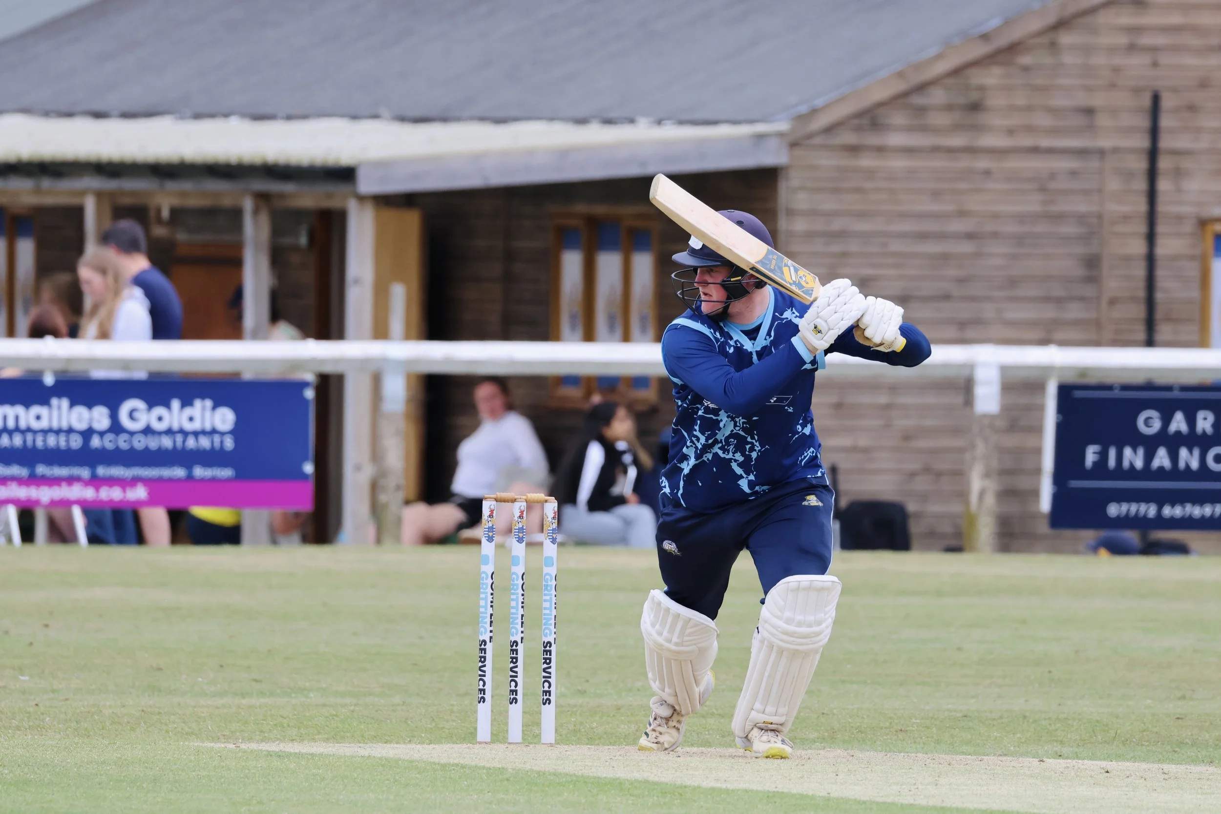 Cricketer in blue uniform prepares to hit the cricket ball during a match on a grassy field, with wooden buildings and spectators in the background.