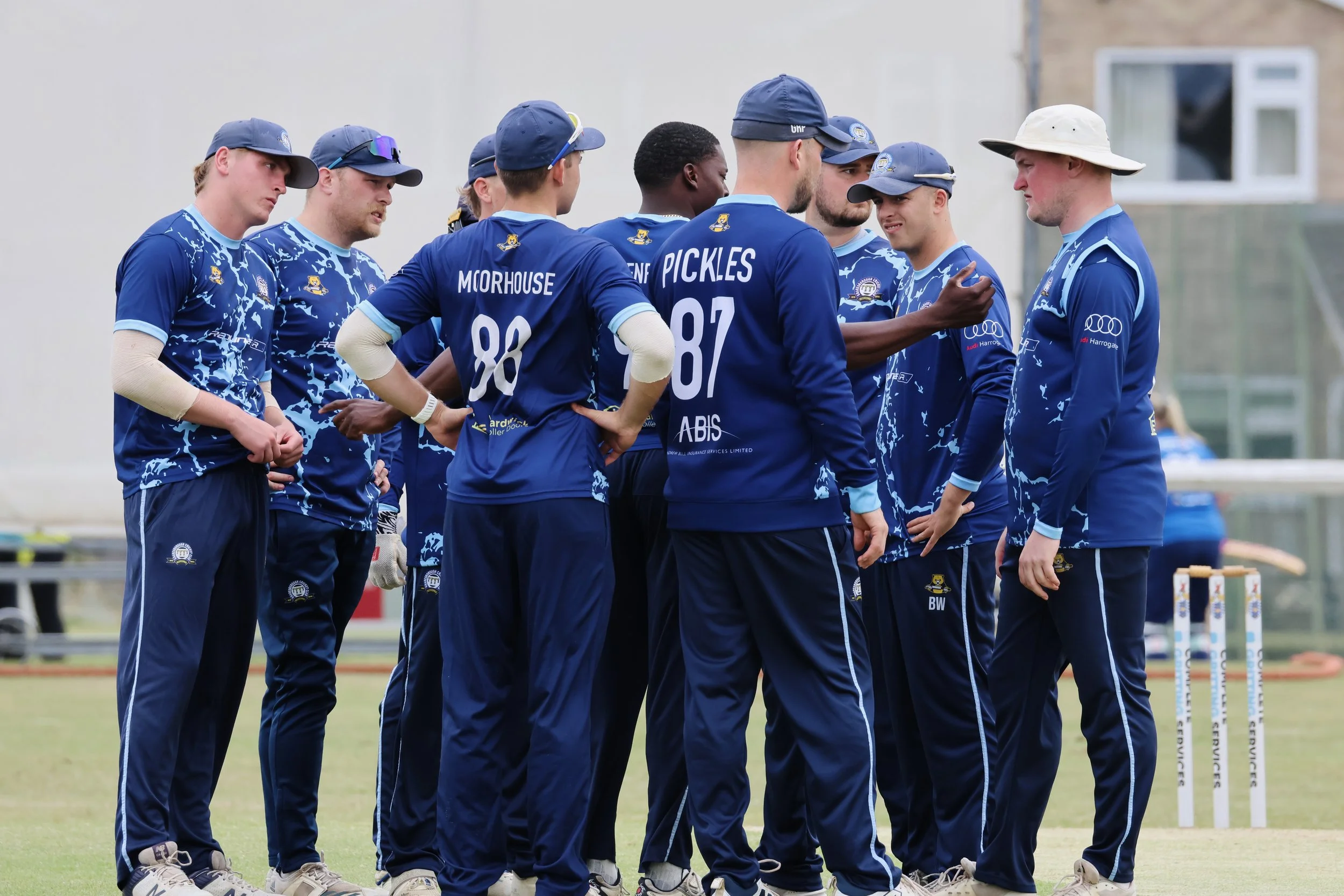 Cricket players in blue uniforms and caps standing on the field in a huddle.