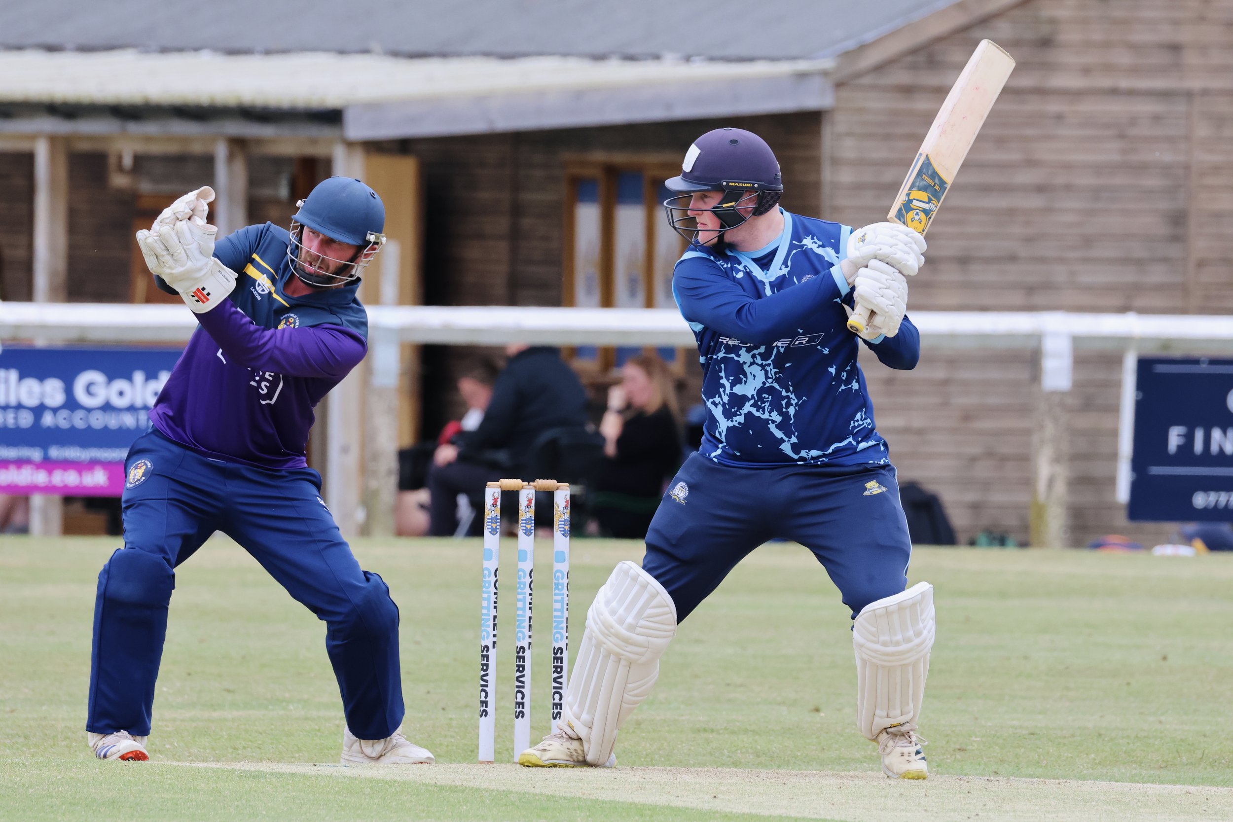 Two cricket players wearing helmets, gloves, and uniforms playing cricket on a field. One player is holding a bat, preparing to hit the ball, while the other appears to be in a defensive stance near the stumps.