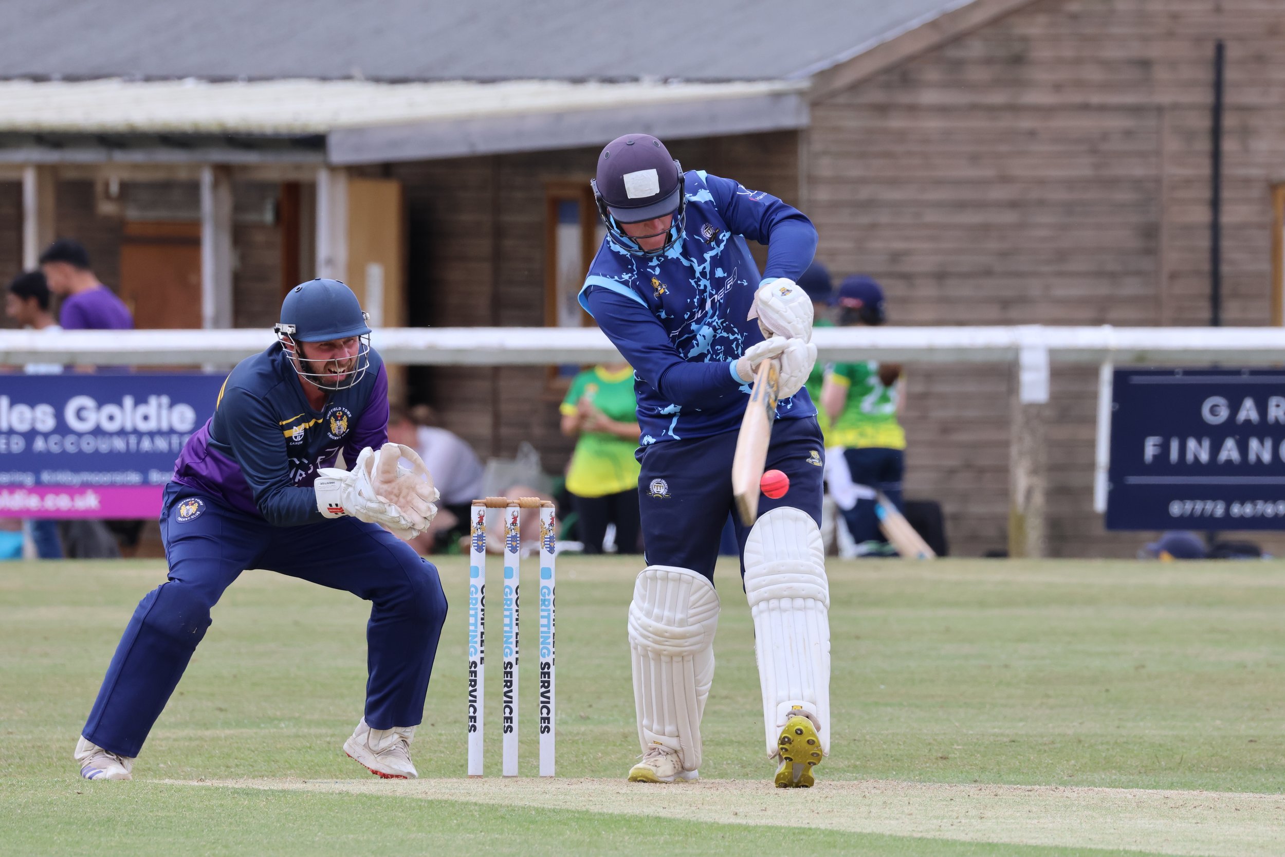 A cricket player in blue uniform hitting a ball while another player in purple and blue crouches behind the wickets on a cricket field.