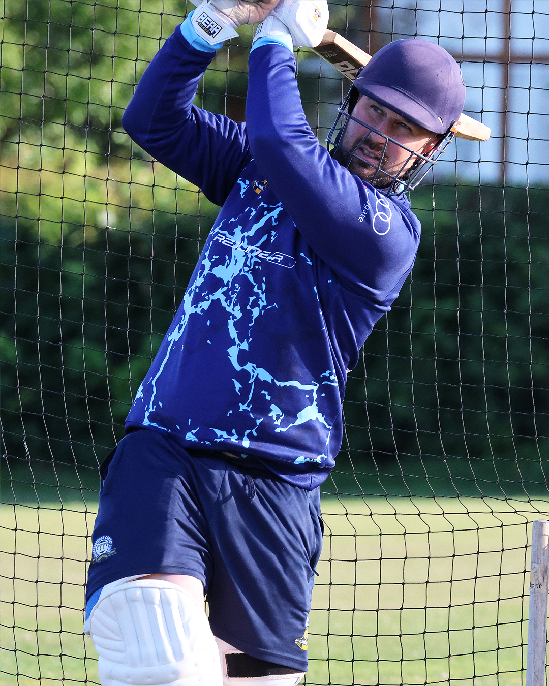 A man practicing baseball outdoors, wearing a purple helmet and blue sports jersey, holding a bat, standing in front of a safety net with green trees in the background.