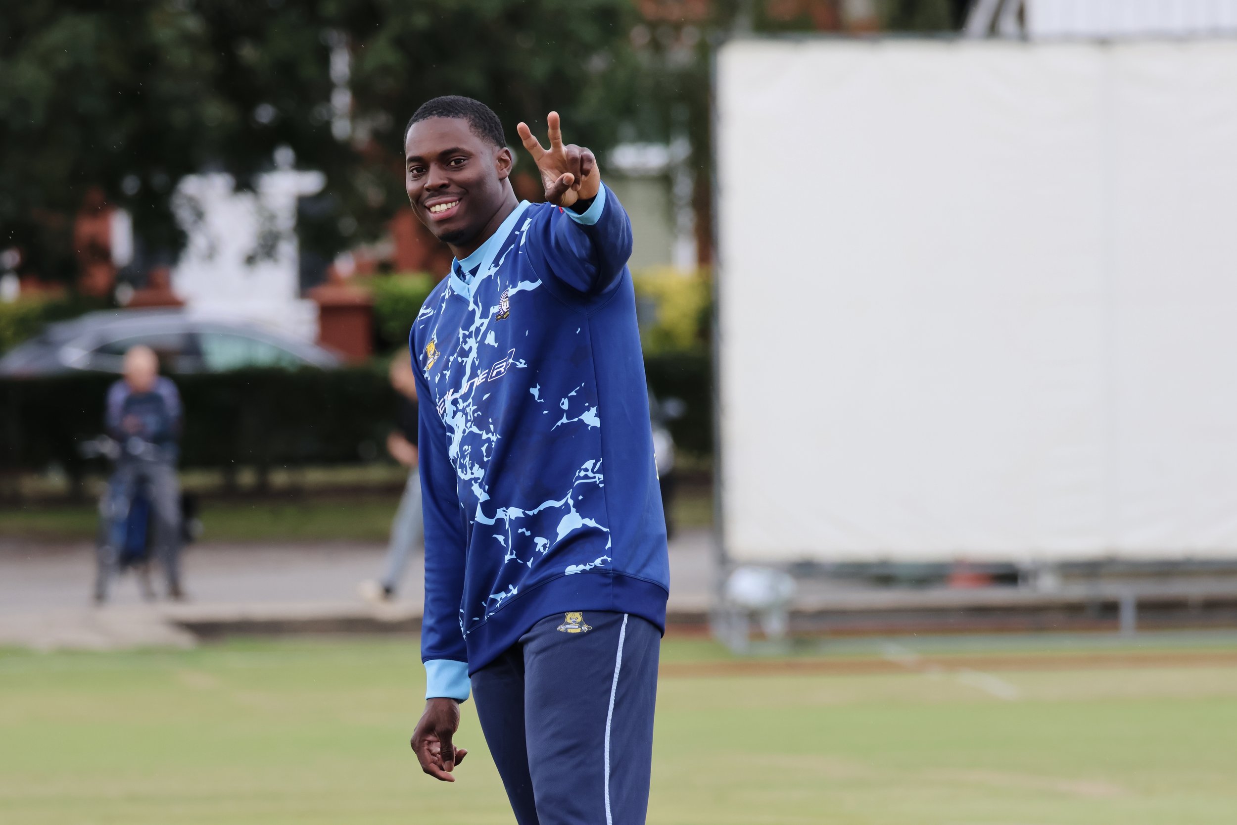 Young man in a blue sports jersey smiling and waving outdoors on a sports field.