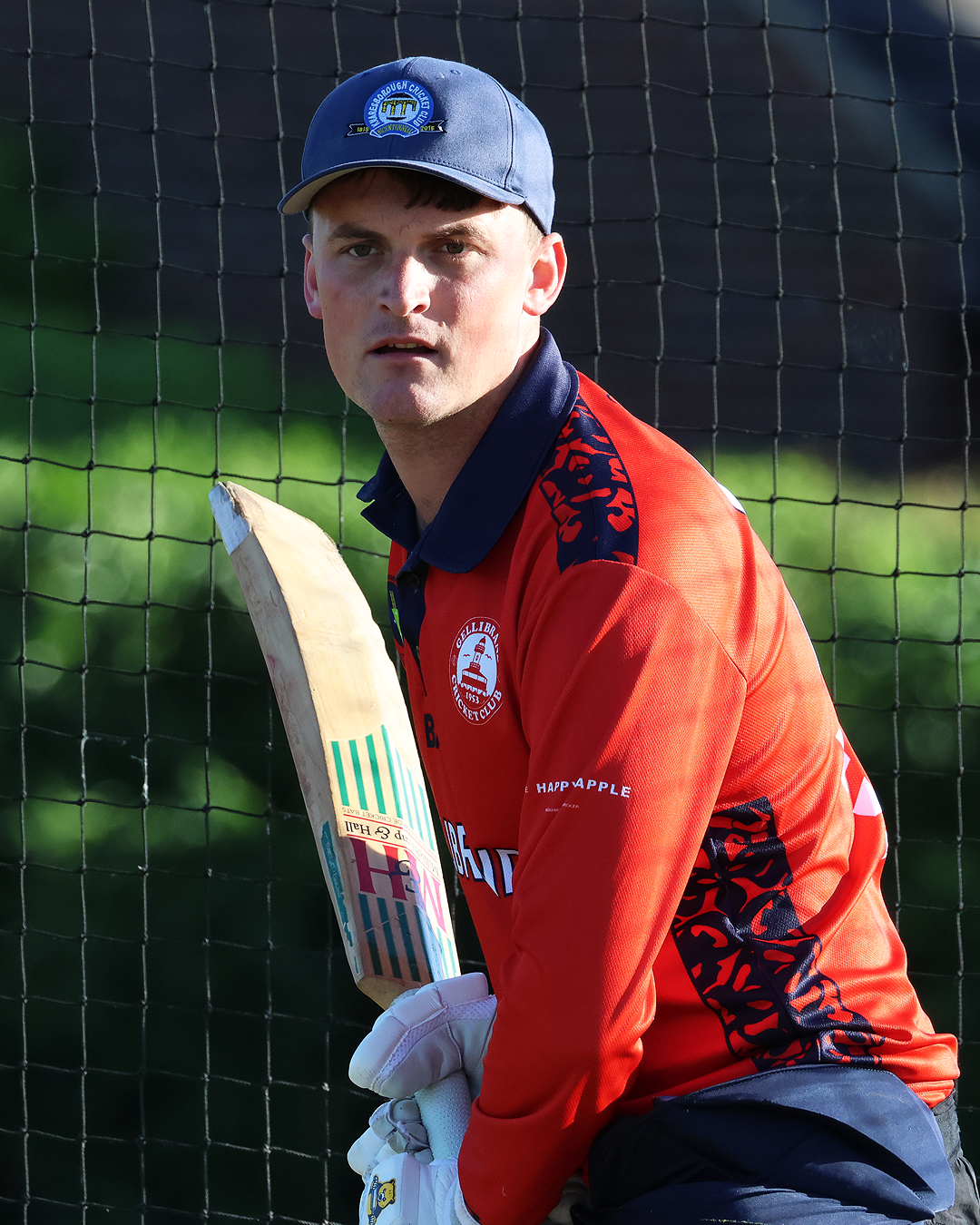 Cricket player in red uniform and blue cap holding a cricket bat, standing near a net with Green background.