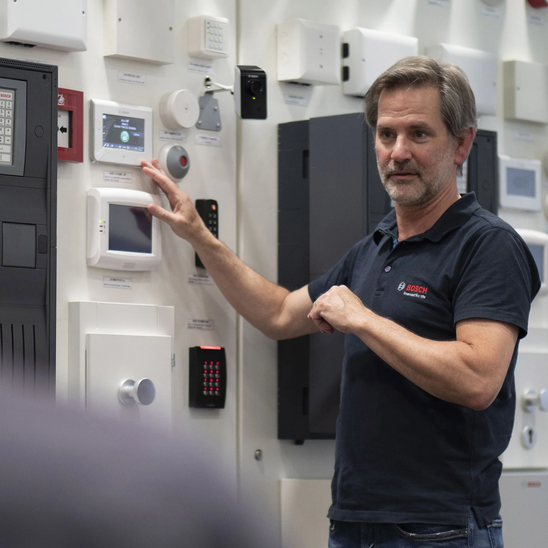 A man with gray hair and beard, wearing a black polo shirt with a Bosch logo, pointing at a white control panel on a wall of various electrical devices and security equipment.