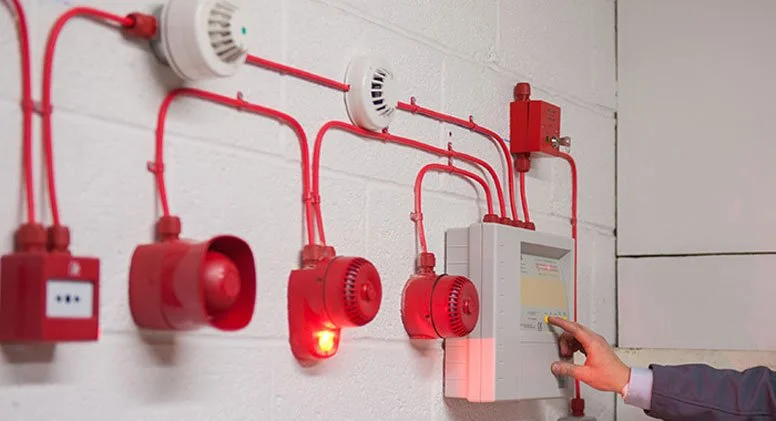 Person operating a fire alarm control panel on a white brick wall with red pipes and smoke detectors.