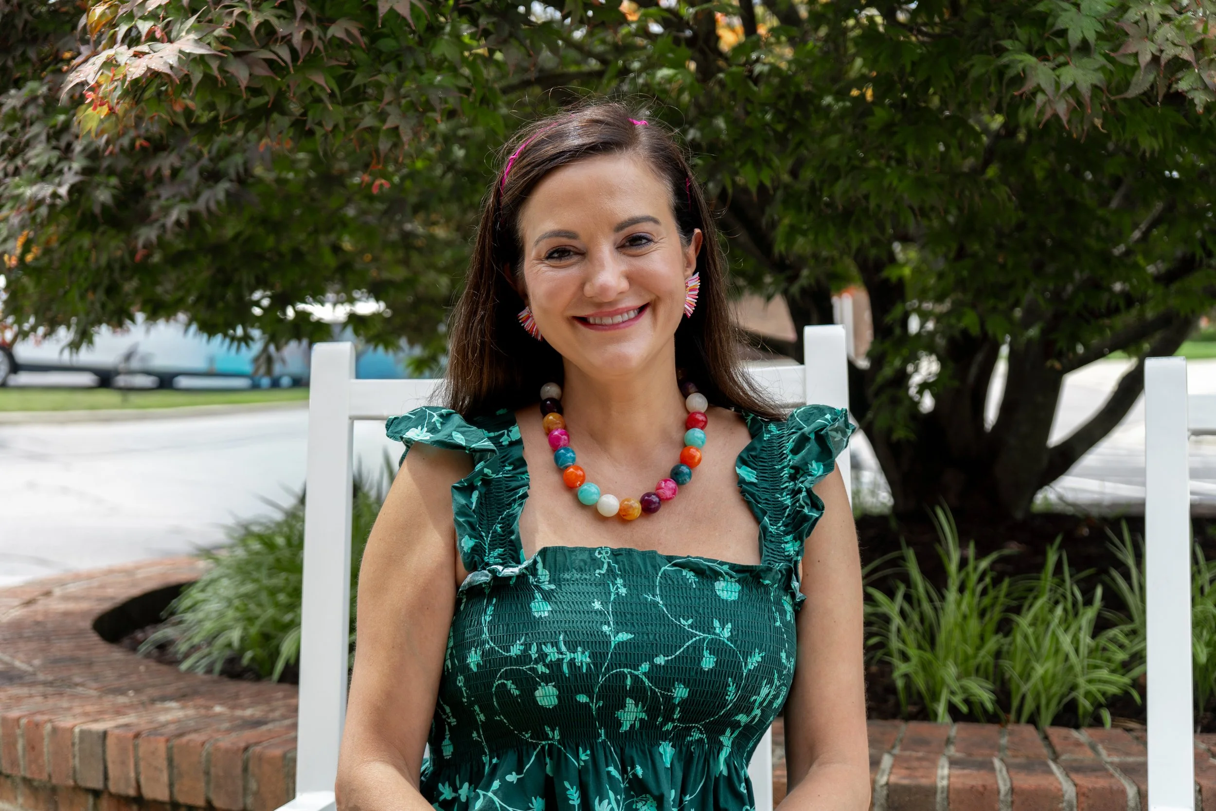 A woman with long brown hair is smiling and sitting outdoors in front of a leafy tree. She is wearing a green dress with a floral pattern, a colorful bead necklace, and colorful earrings. There is a white chair behind her.