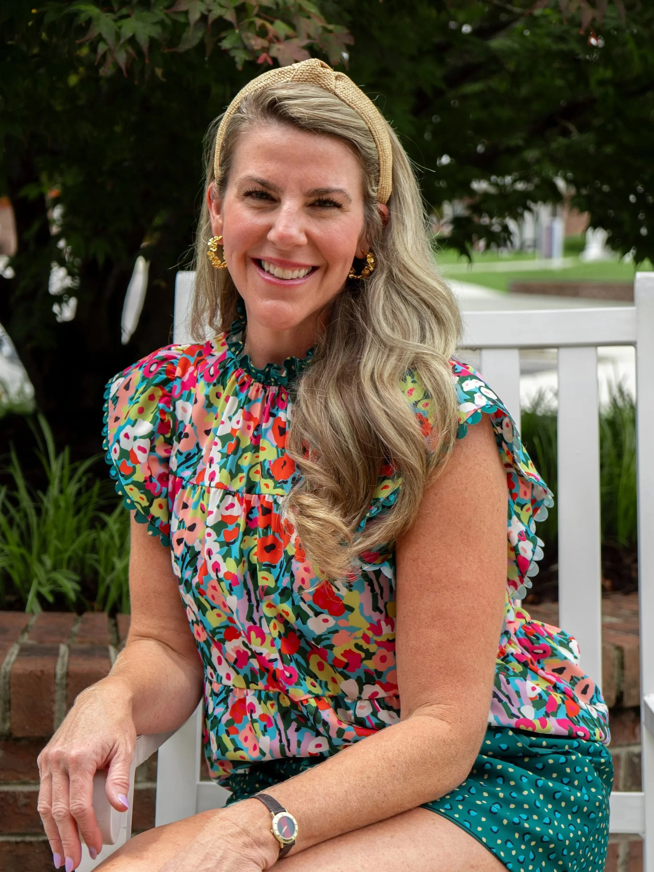A woman with long blonde hair, wearing a colorful, patterned dress, a beige headband, and gold earrings, sitting outdoors on a white chair, smiling at the camera, with green trees and a brick wall in the background.