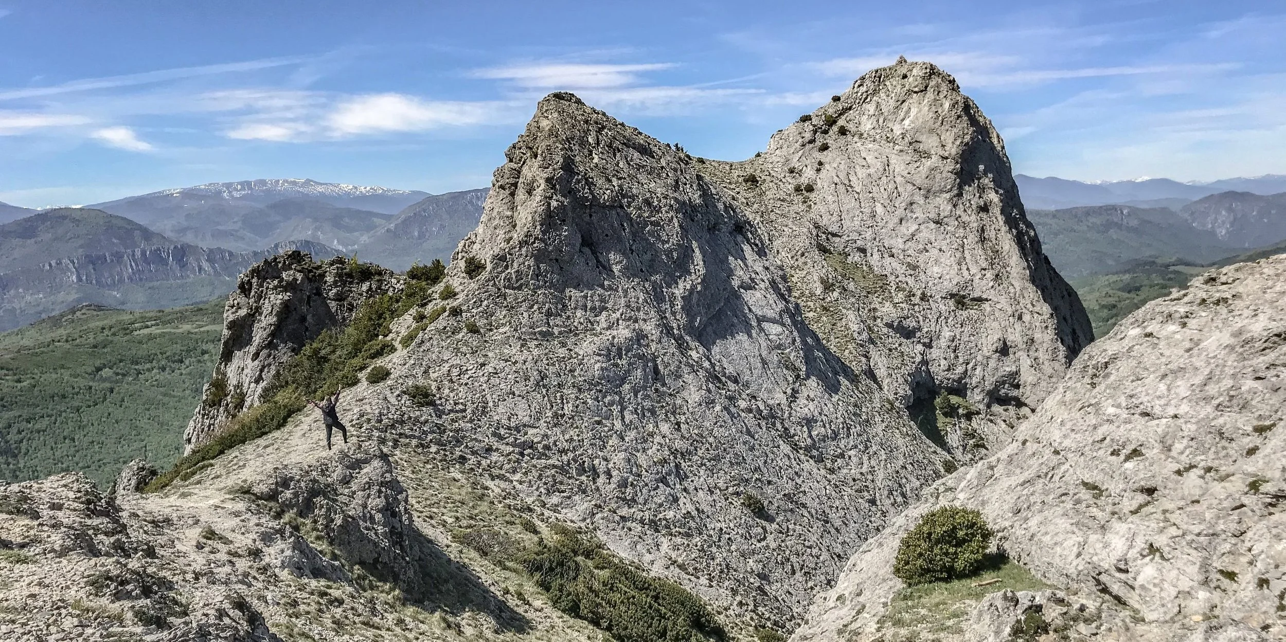 A person climbing and standing on large rocky mountain terrain with a mountain range and blue sky in the background.