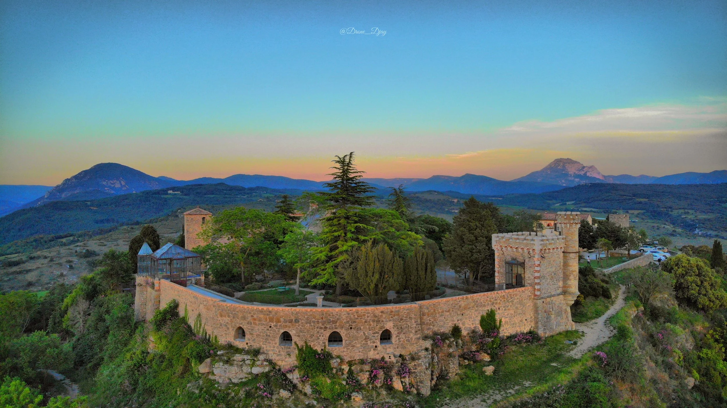 Scenic view of a castle with stone walls and towers surrounded by greenery, with mountains in the background and a colorful sky at dusk.