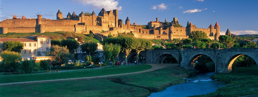 A medieval castle with multiple towers on a hill, with a stone bridge over a river in the foreground and a park with trees and a path.