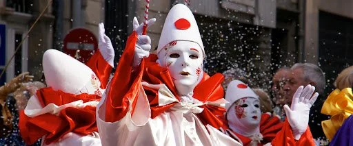 People dressed as white-faced masks and red costumes participating in a parade, with one person holding a stick with decorations, and snow-like confetti in the air.