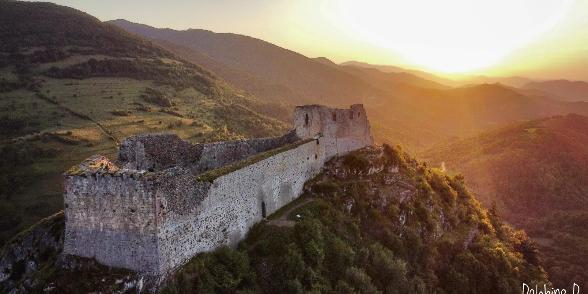 Ancient stone castle ruins on a hilltop at sunset, overlooking green mountains and valleys.
