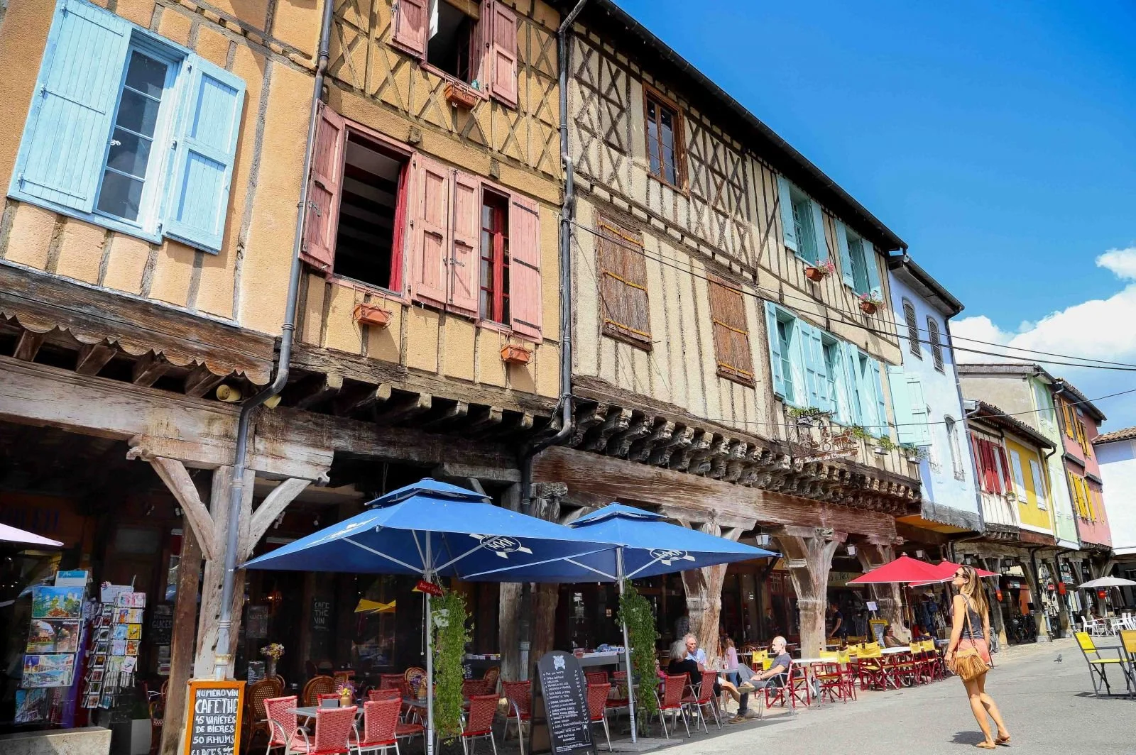 Colorful storefronts with outdoor seating, umbrellas, and pedestrians under a clear blue sky in a lively street scene.