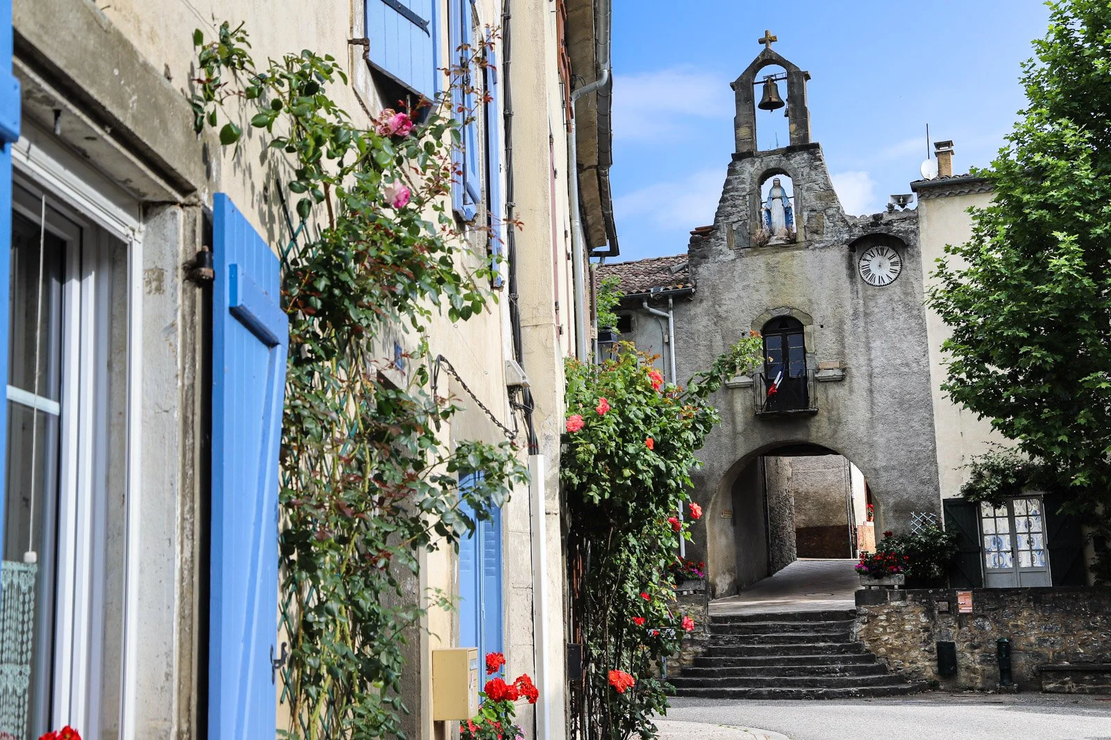 A narrow street in a small village with flowers and buildings with blue shutters, leading to a stone church with a clock, statue, and bell tower, under a blue sky.