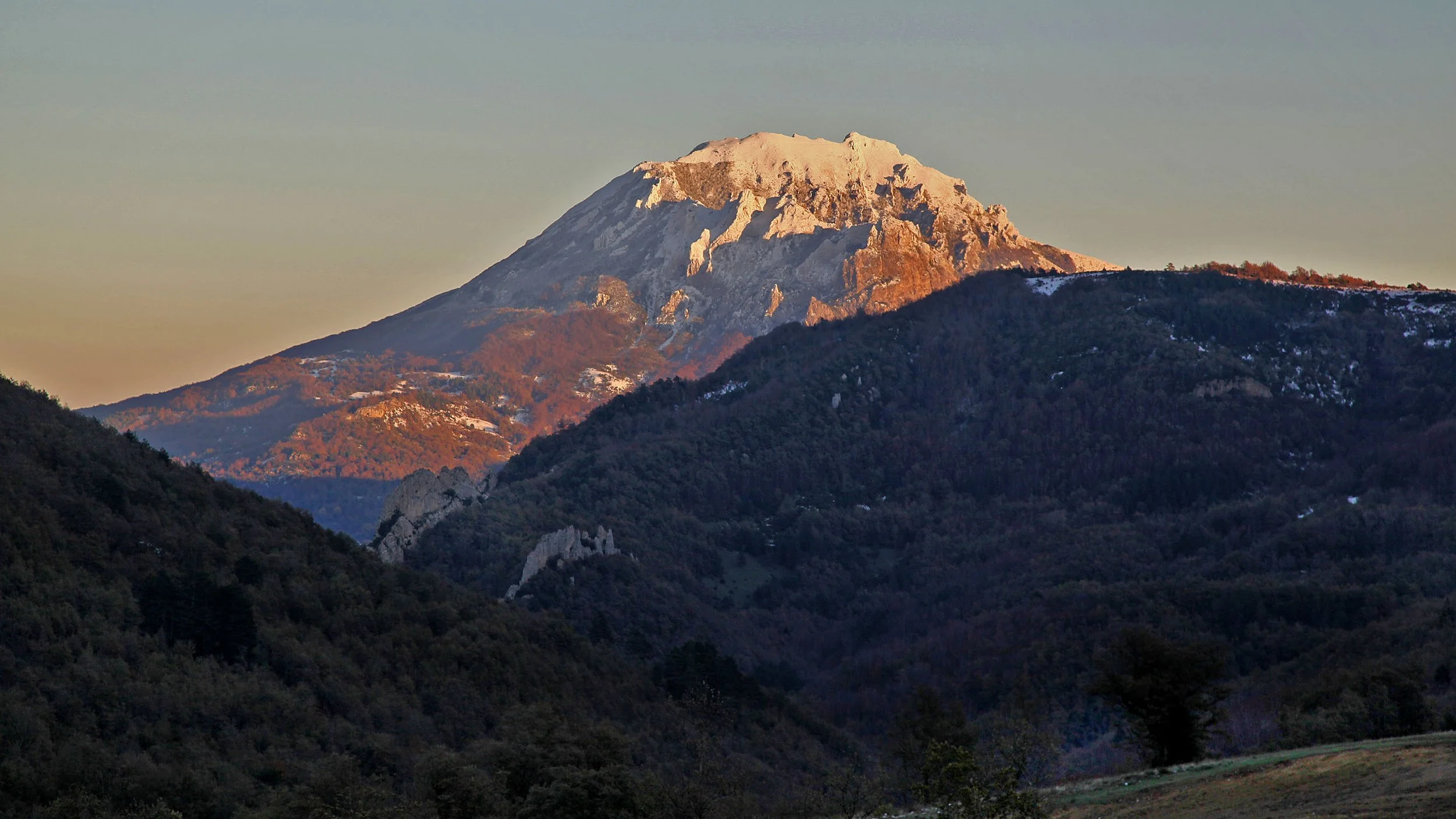 A mountain covered in snow, with a forested valley below during sunset.