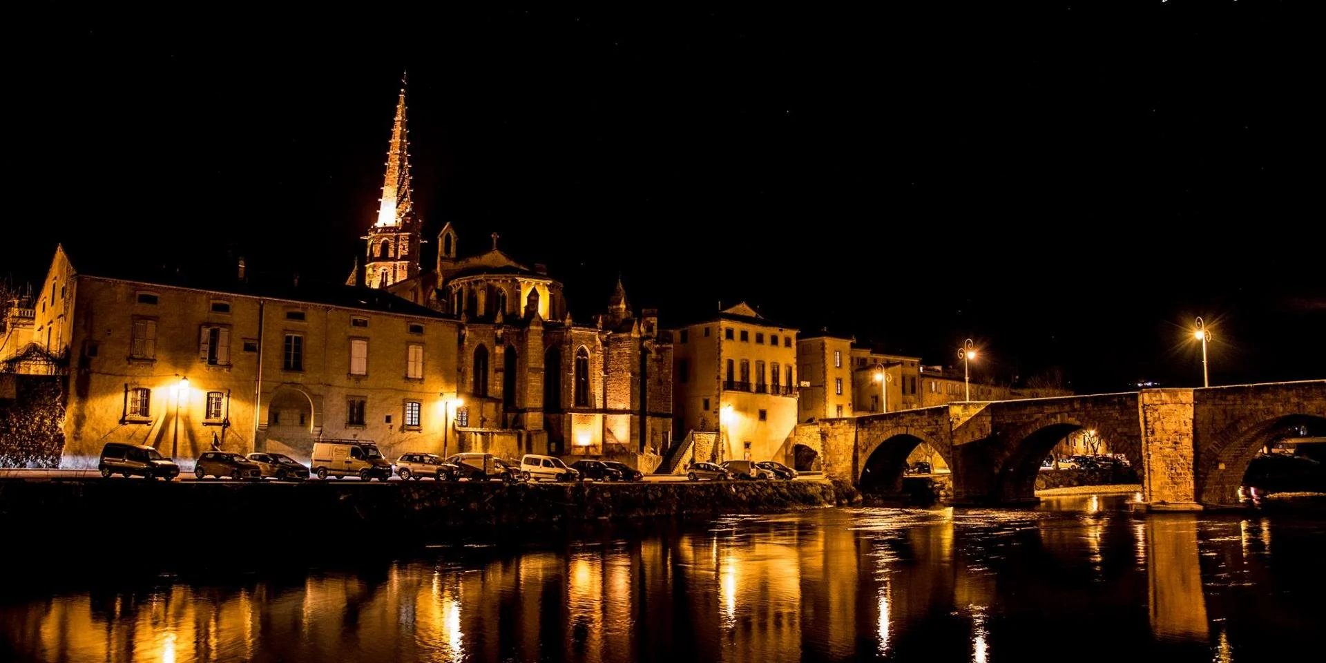 Night view of historic buildings and a bridge over a river, illuminated by streetlights and the buildings' lights, with a clear starry sky above.